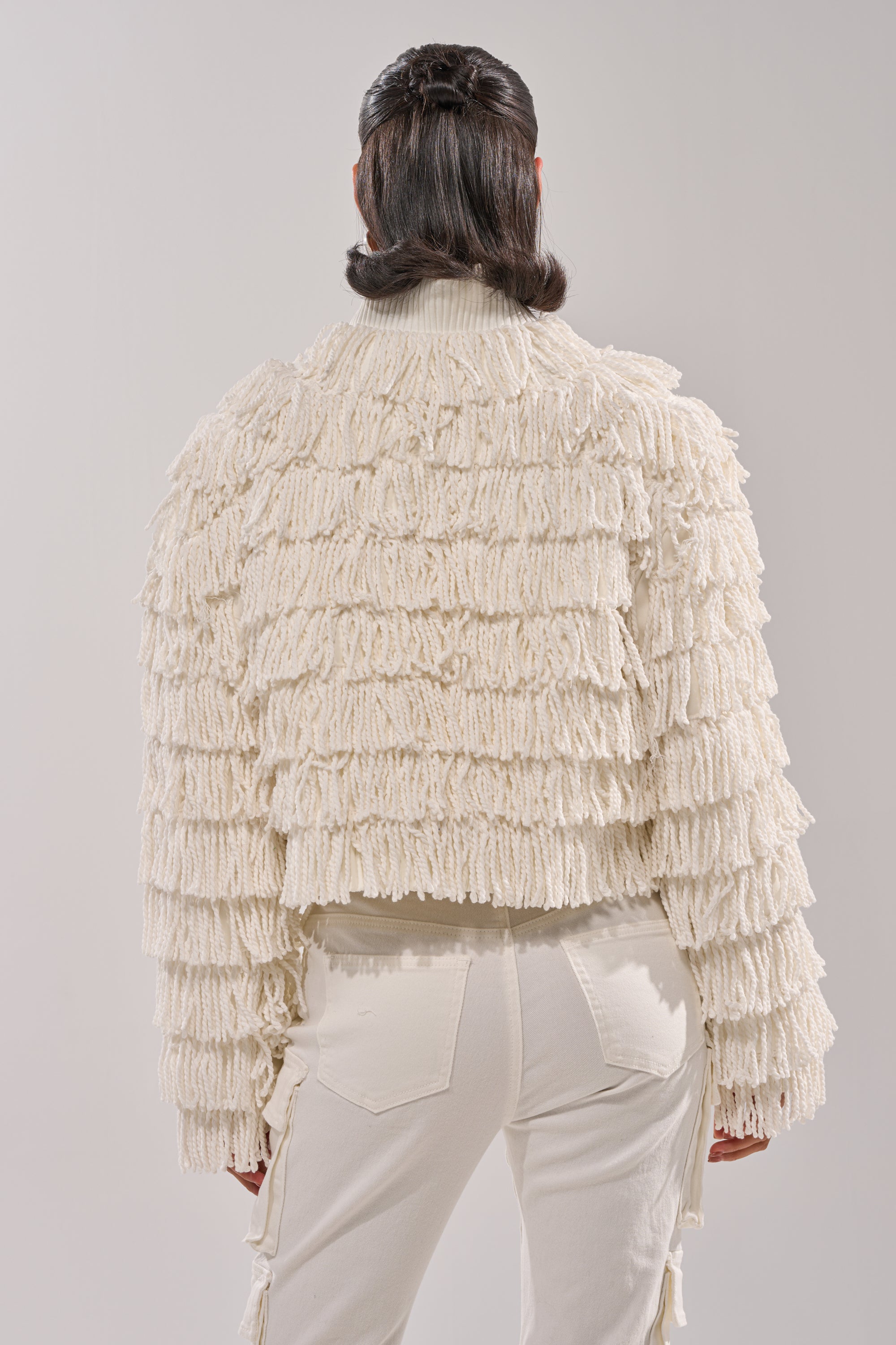 A person faces away from the camera, wearing the FREYA FRINGE BOMBER— a cream-colored jacket with a shaggy fringe finish— paired with matching light pants against a neutral background.