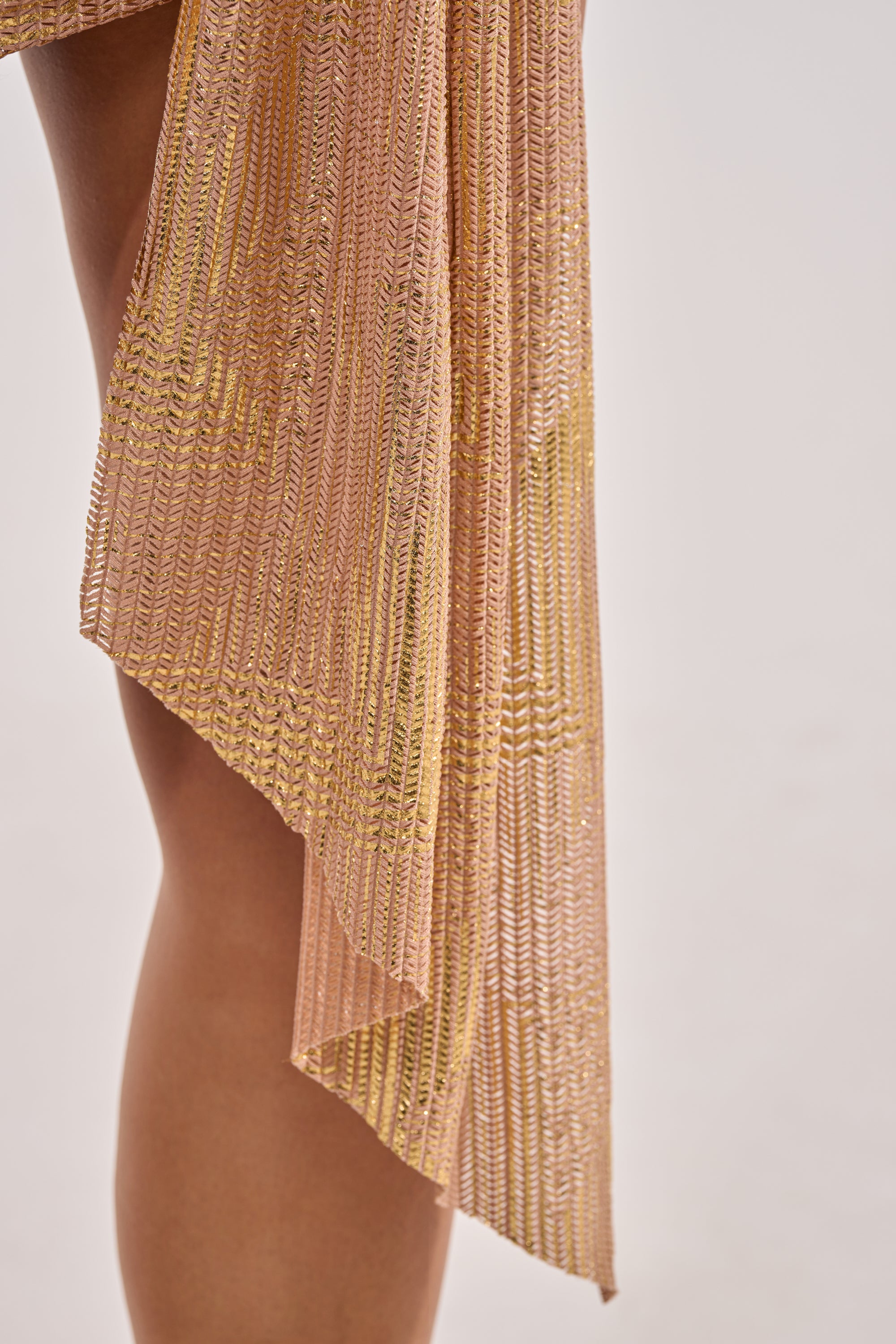 Close-up of a person’s leg in the DREAM GIRL WOMEN MINI DRESS IN GOLD—sheer, gold and pink pleated party dress with an asymmetrical hem, shown against a plain light background. 6