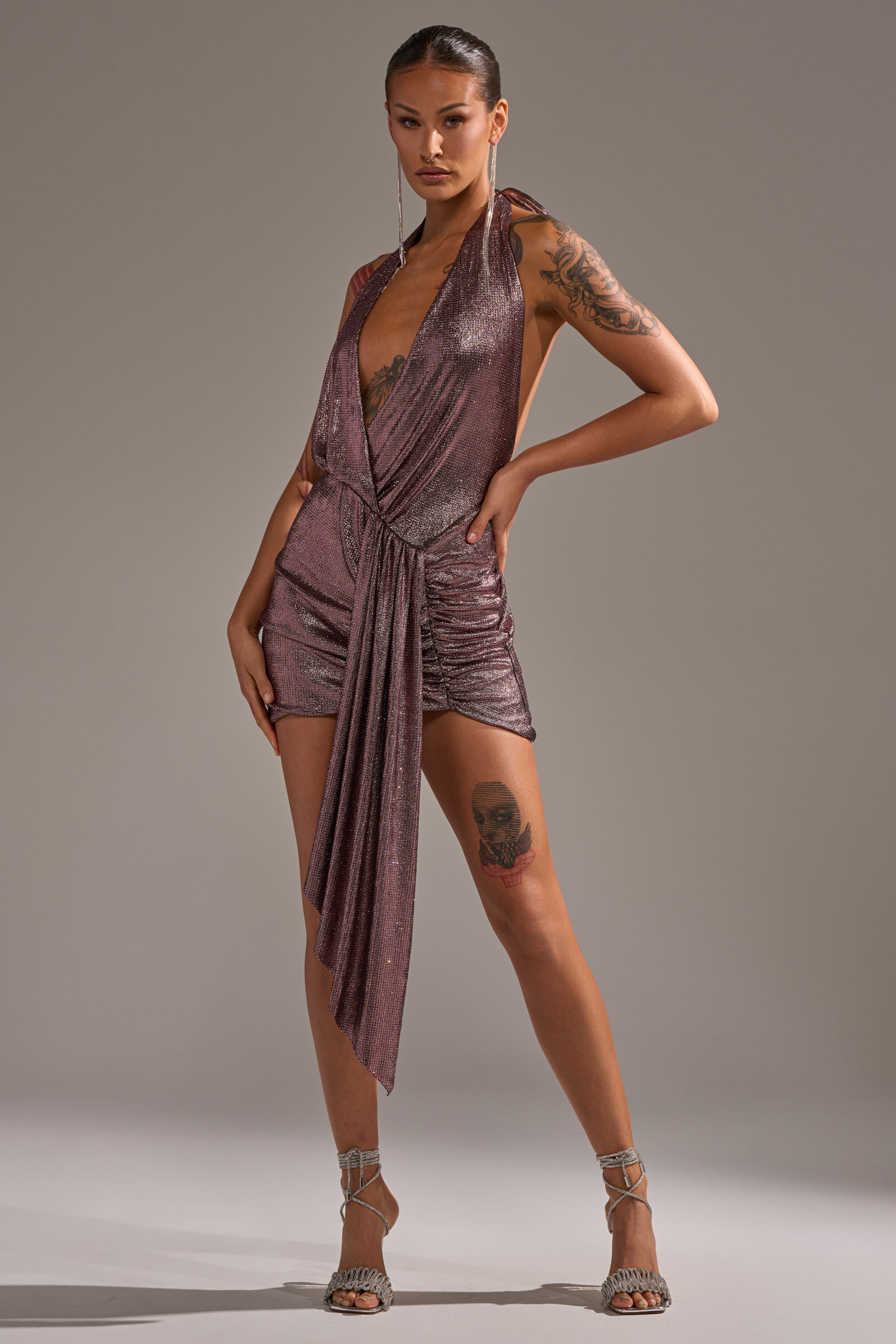 A woman poses confidently in the DREAM GIRL RHINESTONE MINI DRESS IN PINK, a sleeveless mini dress with draped detailing and a deep V-neck. She shows off her tattoos, wears strappy heels, and has her hair slicked back. 9