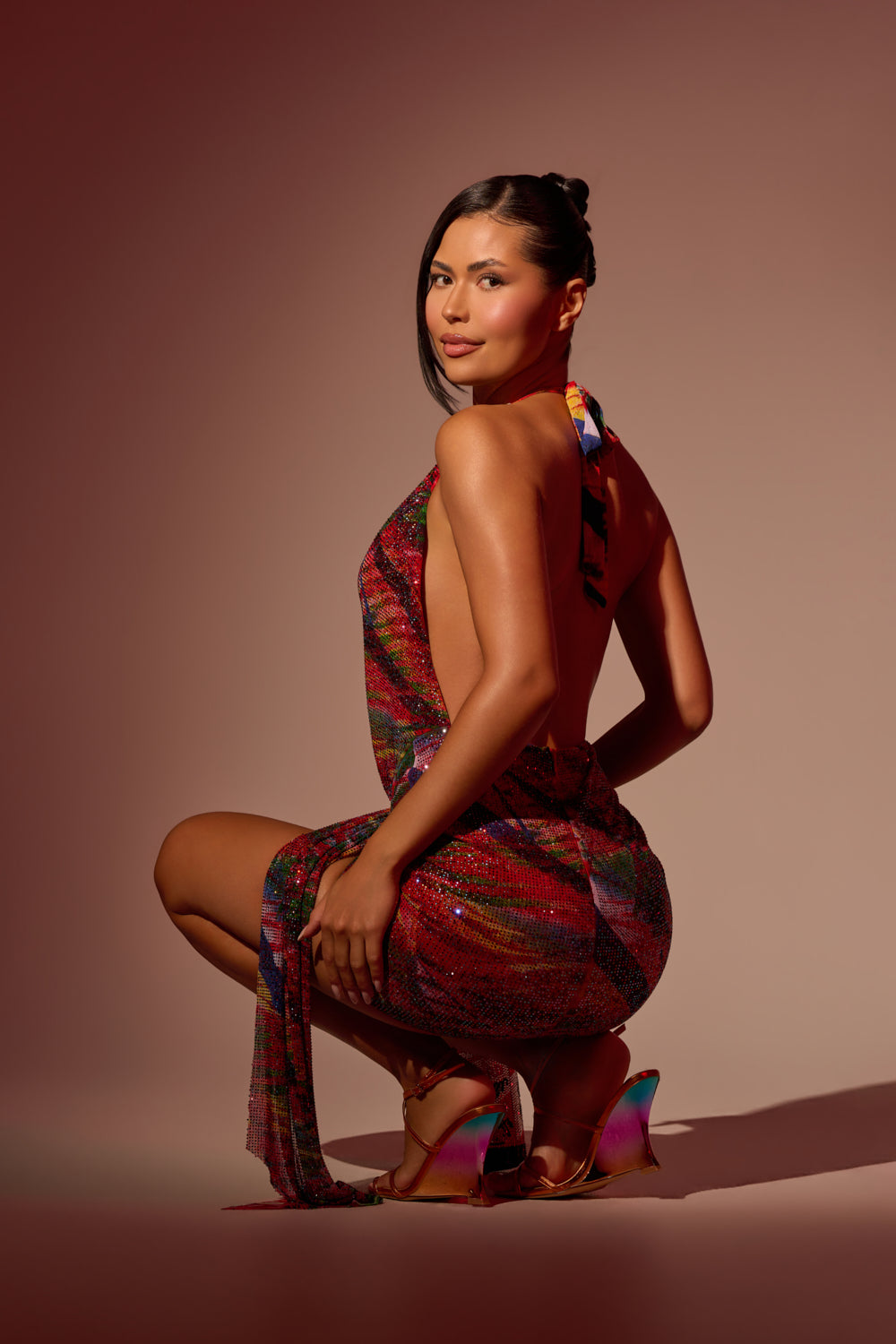 A woman with dark hair in a sleek updo squats against a neutral gradient background, wearing the FEVER DREAM RHINESTONE MINI DRESS and shiny high-heeled sandals. 3