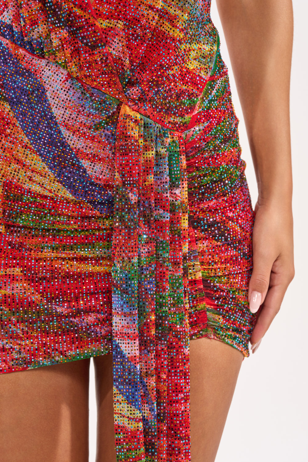 A close-up shows someone in the FEVER DREAM RHINESTONE MINI DRESS—a sparkly red halter mini with ruched side and tie detail. Their hand highlights the vibrant textured fabric, making this dress perfect for any festive occasion. 8