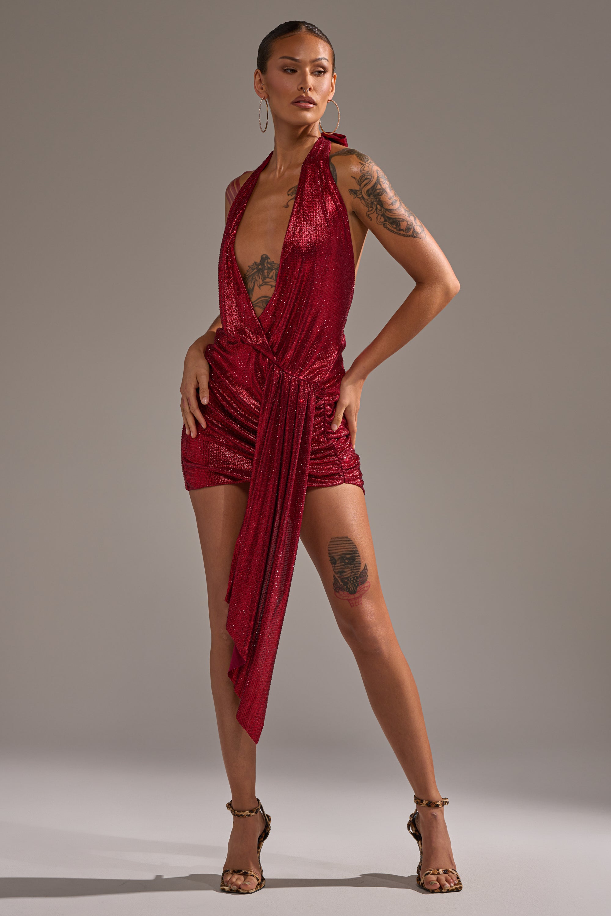 A woman models the DREAM GIRL RHINESTONE DRESS IN RED, a sleeveless, shimmering wrap dress with a plunging neckline and asymmetrical draping. She accessorizes with hoop earrings, strappy heels, and displays tattoos on her arm and leg. 9