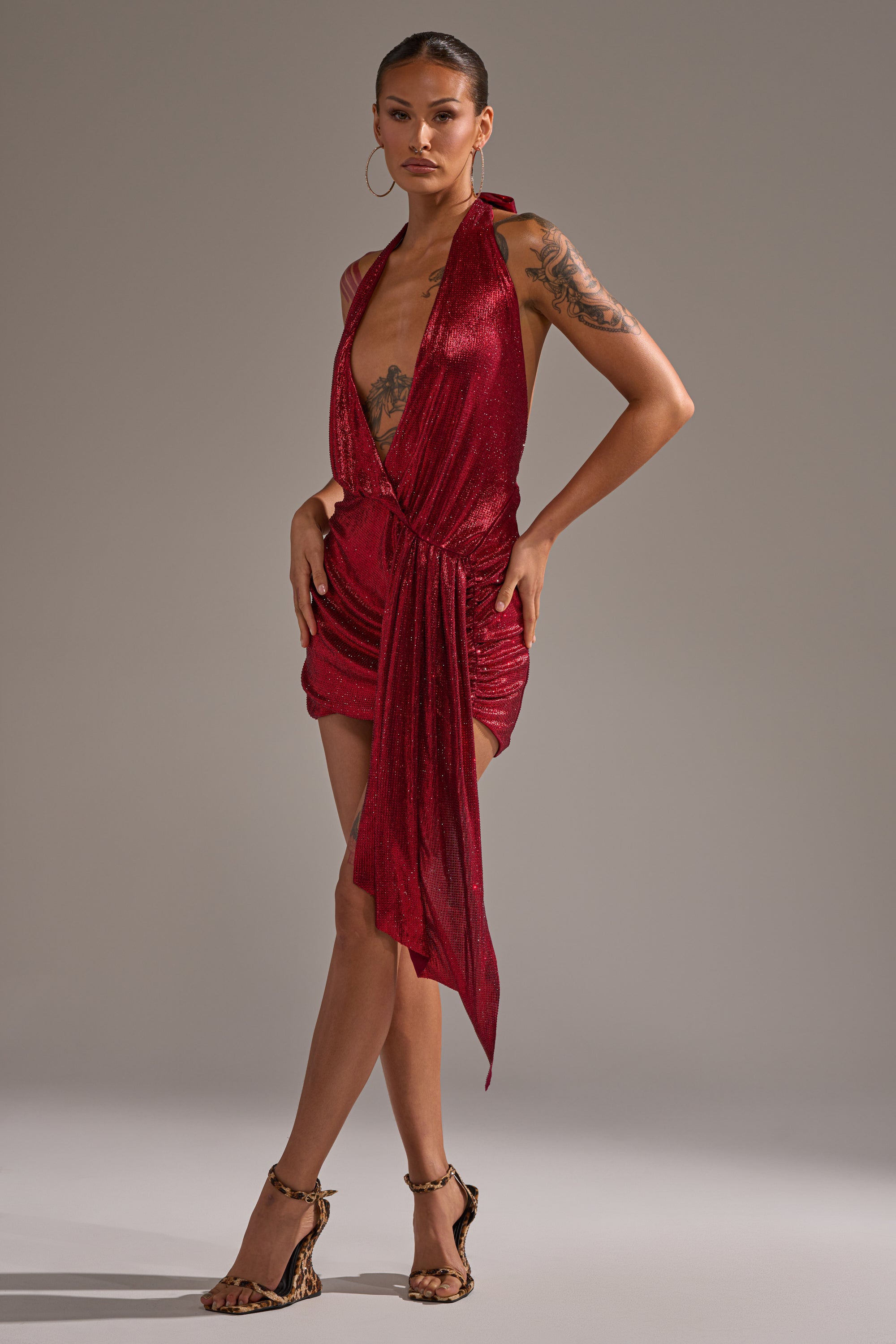 A woman poses against a plain gray background in the DREAM GIRL RHINESTONE DRESS IN RED—a shimmering, sleeveless mini dress with a plunging neckline and draped detailing—styled with hoop earrings and animal print heels. 10