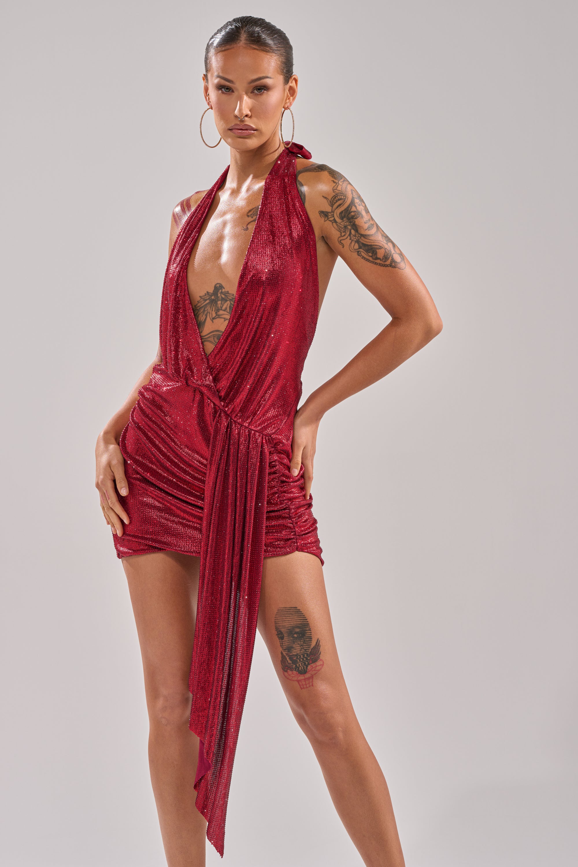 Wearing the DREAM GIRL RHINESTONE DRESS IN RED—a shiny, halter-neck mini dress with a deep V-neckline and draped front detail—a person poses confidently, showcasing tattoos on their arm and thigh against a neutral background. 4
