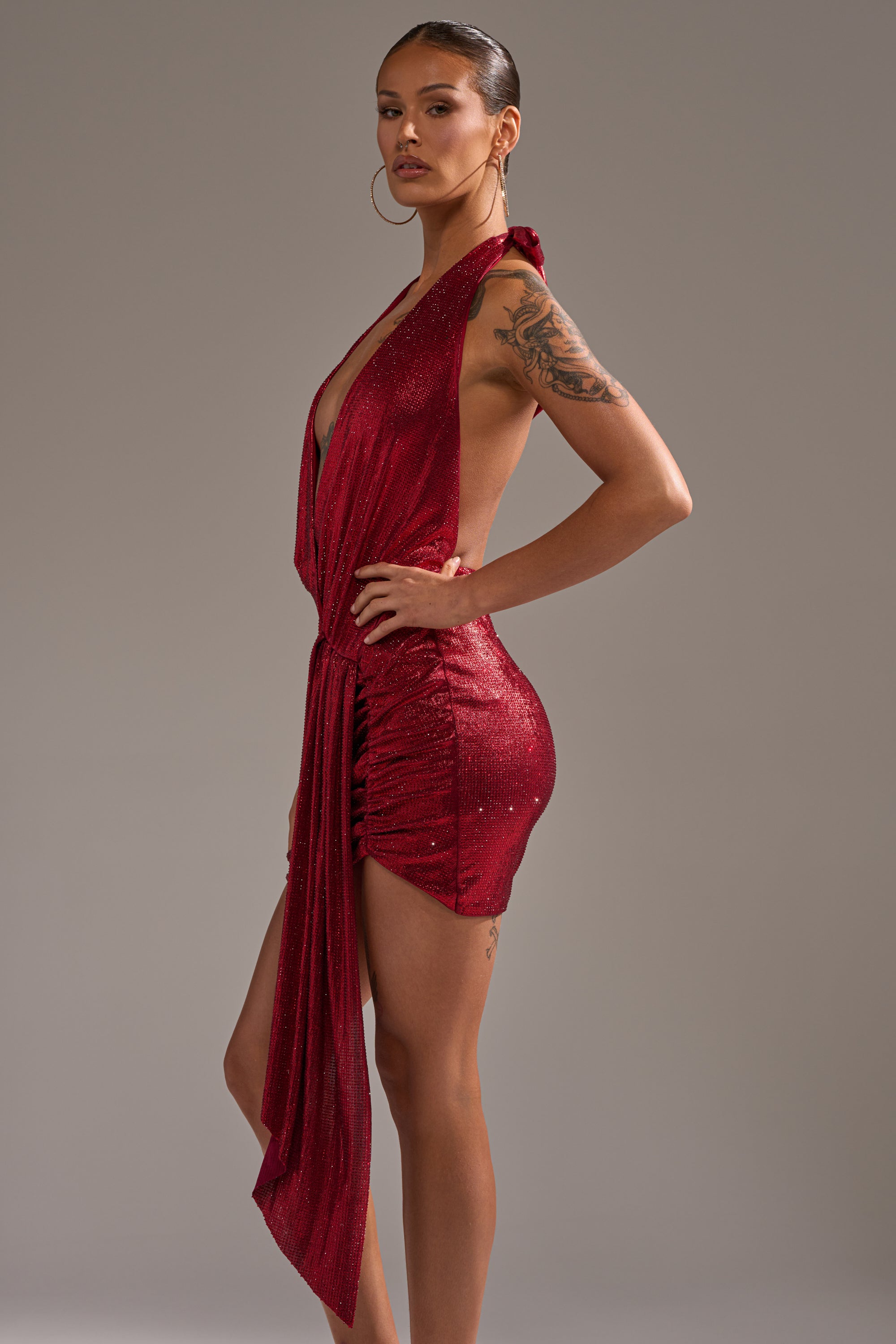 Model showcases the DREAM GIRL RHINESTONE DRESS IN RED—a shimmering, sleeveless dress with a deep neckline and asymmetrical draping. She stands sideways, hand on hip, revealing a striking upper arm tattoo against a plain gray backdrop. 5