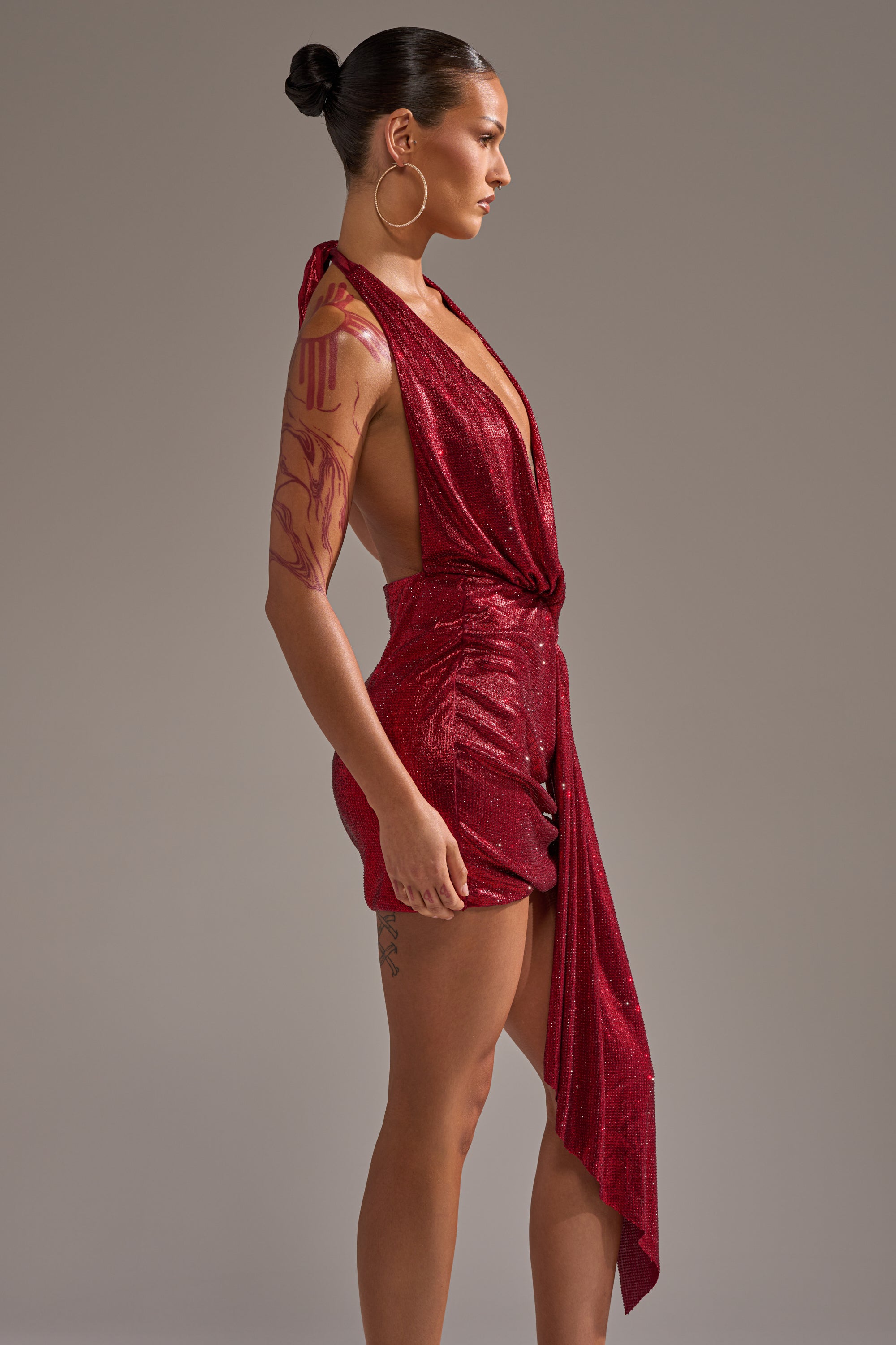 A woman with her hair in a bun, large hoop earrings, and visible arm and thigh tattoos models the DREAM GIRL RHINESTONE DRESS IN RED—a sparkly, sleeveless dress with a deep neckline and draped asymmetrical hem. 6