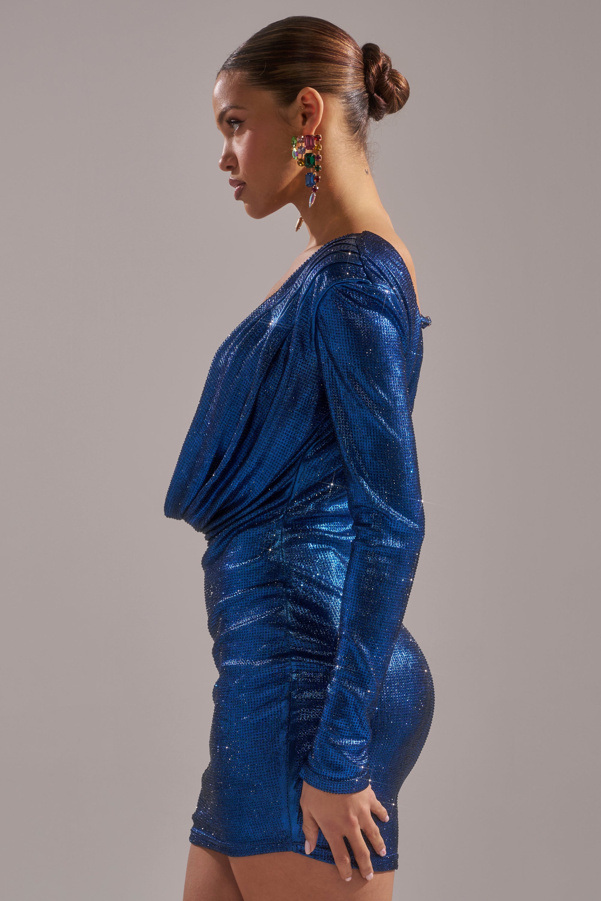 A woman in the TOO HOT TO HANDLE RHINESTONE MINI DRESS IN BLUE stands in profile against a plain backdrop. Her hair is styled in a neat bun, and she completes her look with colorful statement earrings.