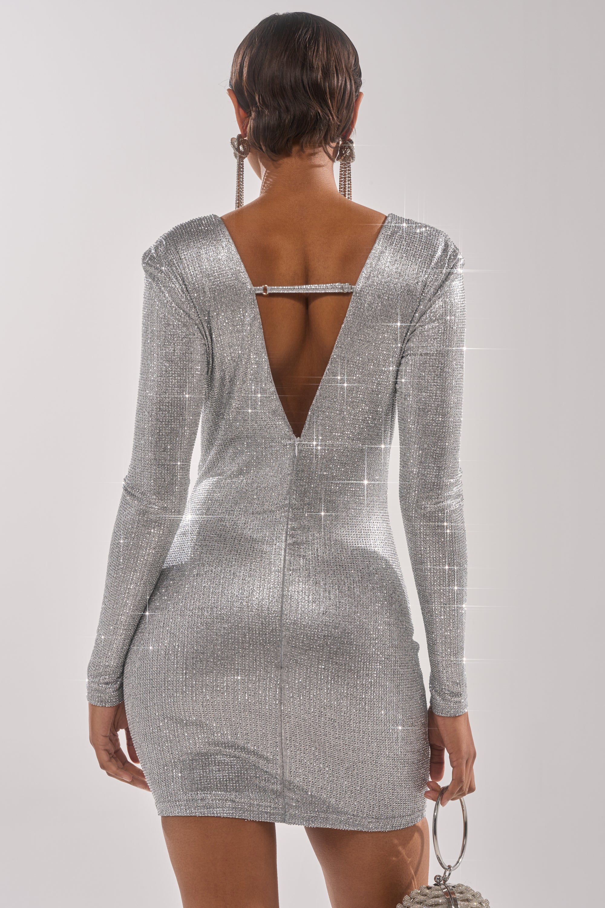 Seen from behind, a person with short hair wears the TOO HOT TO HANDLE RHINESTONE MINI DRESS IN SILVER, featuring long sleeves, an open back with a horizontal strap, and styled with large earrings and a small handbag.