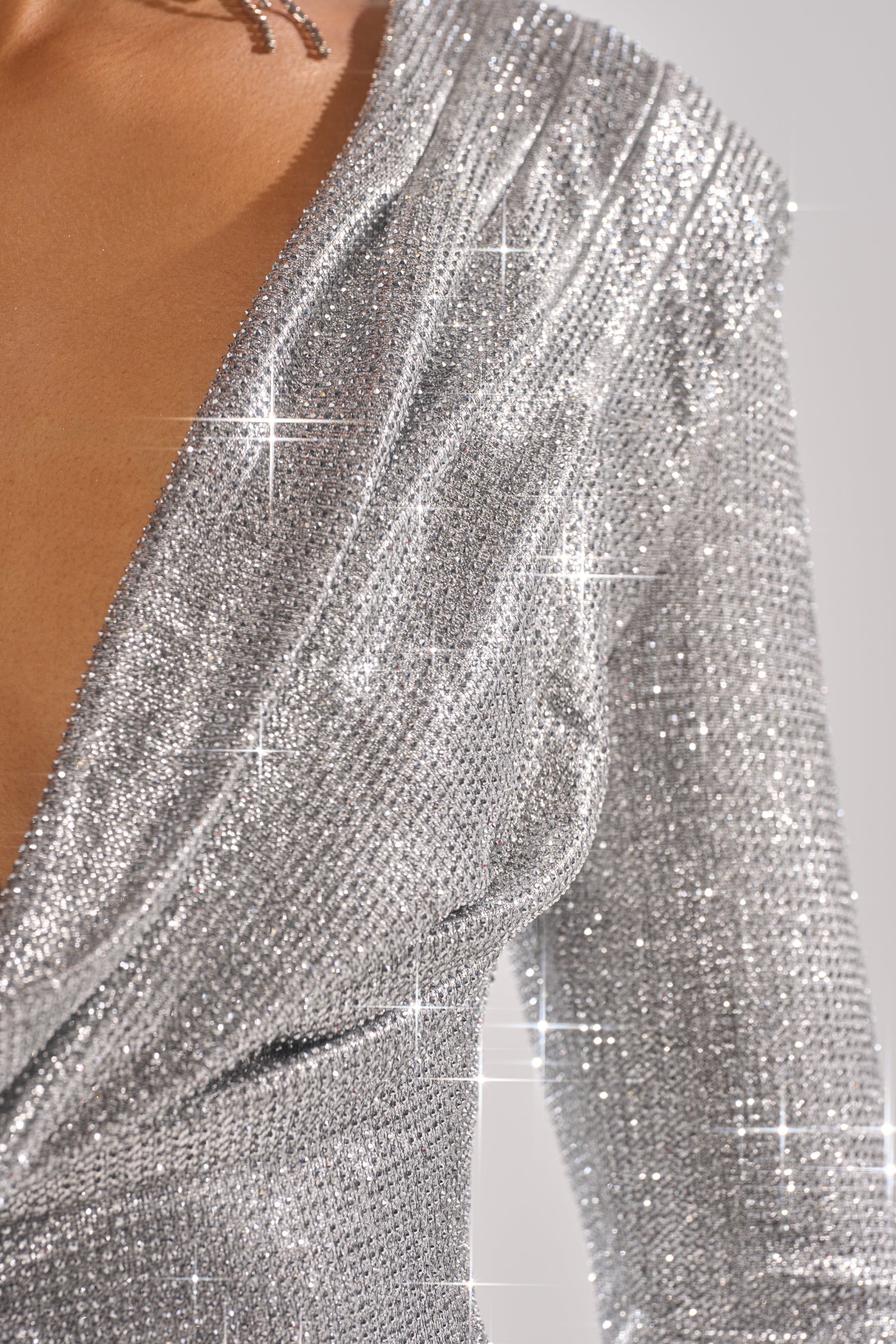 Close-up of a person wearing the TOO HOT TO HANDLE RHINESTONE MINI DRESS IN SILVER, featuring long sleeves and a deep V-neck. The sparkly fabric shimmers under the light, creating a glamorous and elegant look. Only the upper torso is visible.