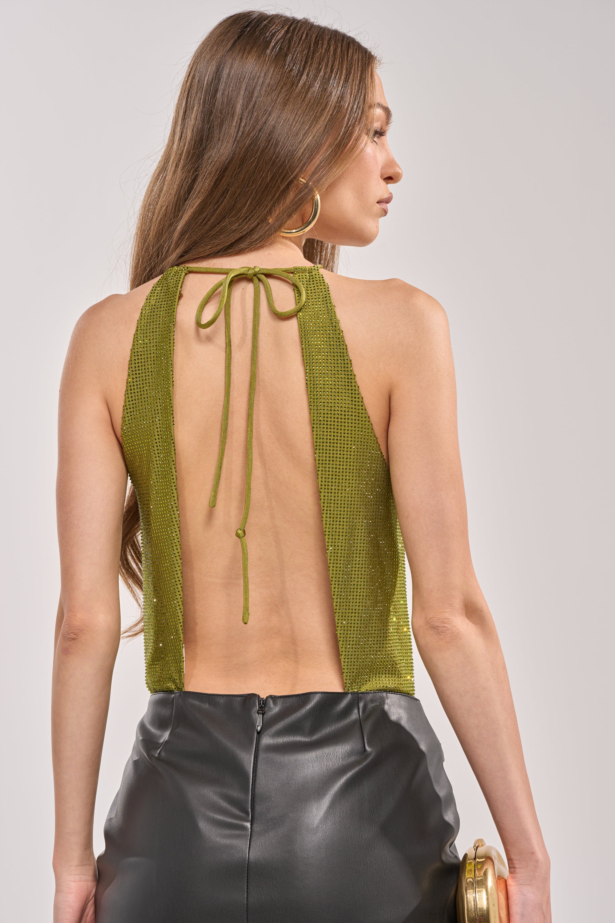 A woman with long brown hair is shown from the back, wearing the SHINING DOWN ON YOU RHINESTONE BODYSUIT IN GREEN, an open-back halter style, paired with a high-waisted black leather skirt and holding a small gold clutch. 3