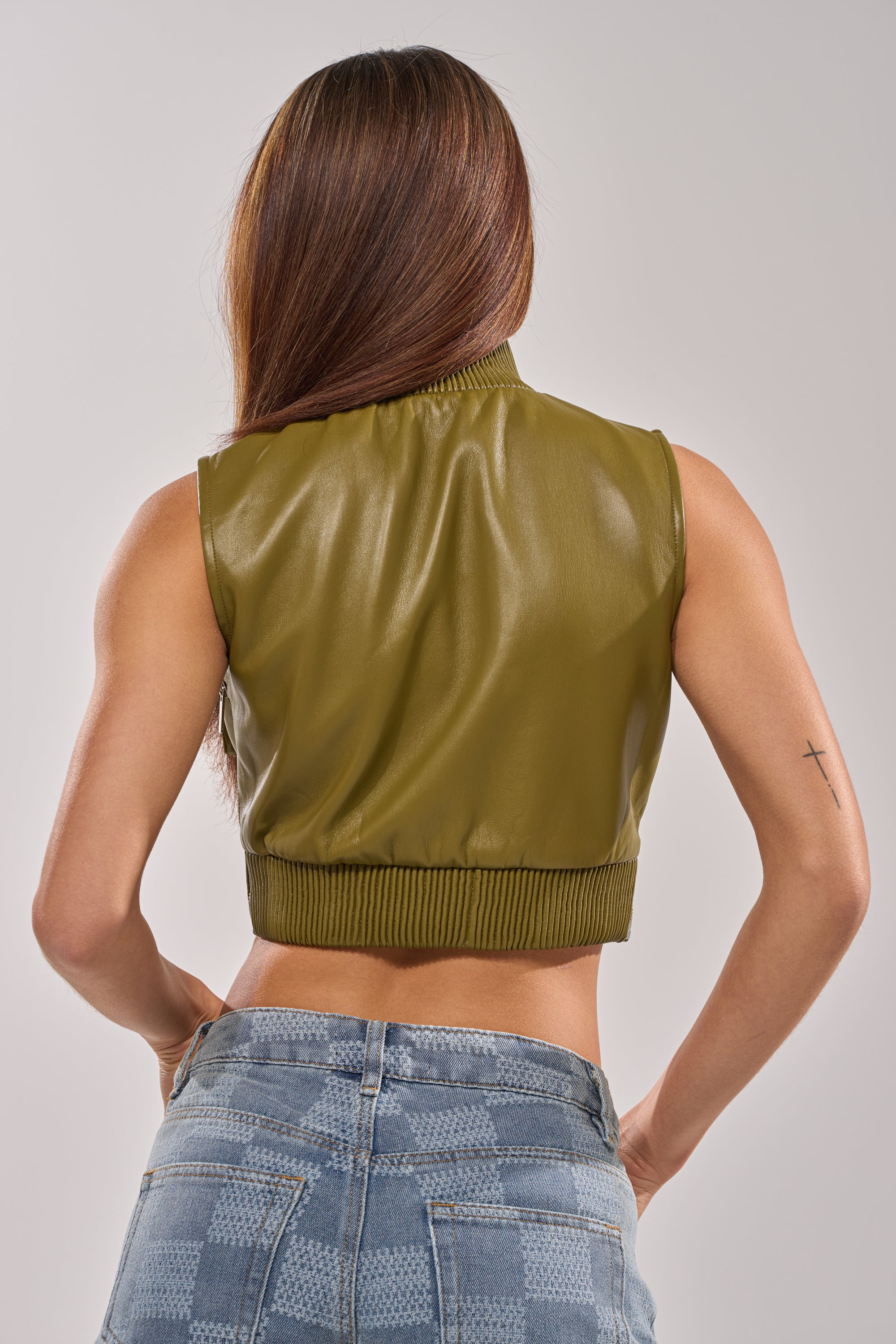 A woman with long brown hair is seen from behind wearing the VANJA MOCK NECK FAUX LEATHER TOP in olive and high-waisted blue checkered jeans, showcasing a small cross tattoo on her left arm.