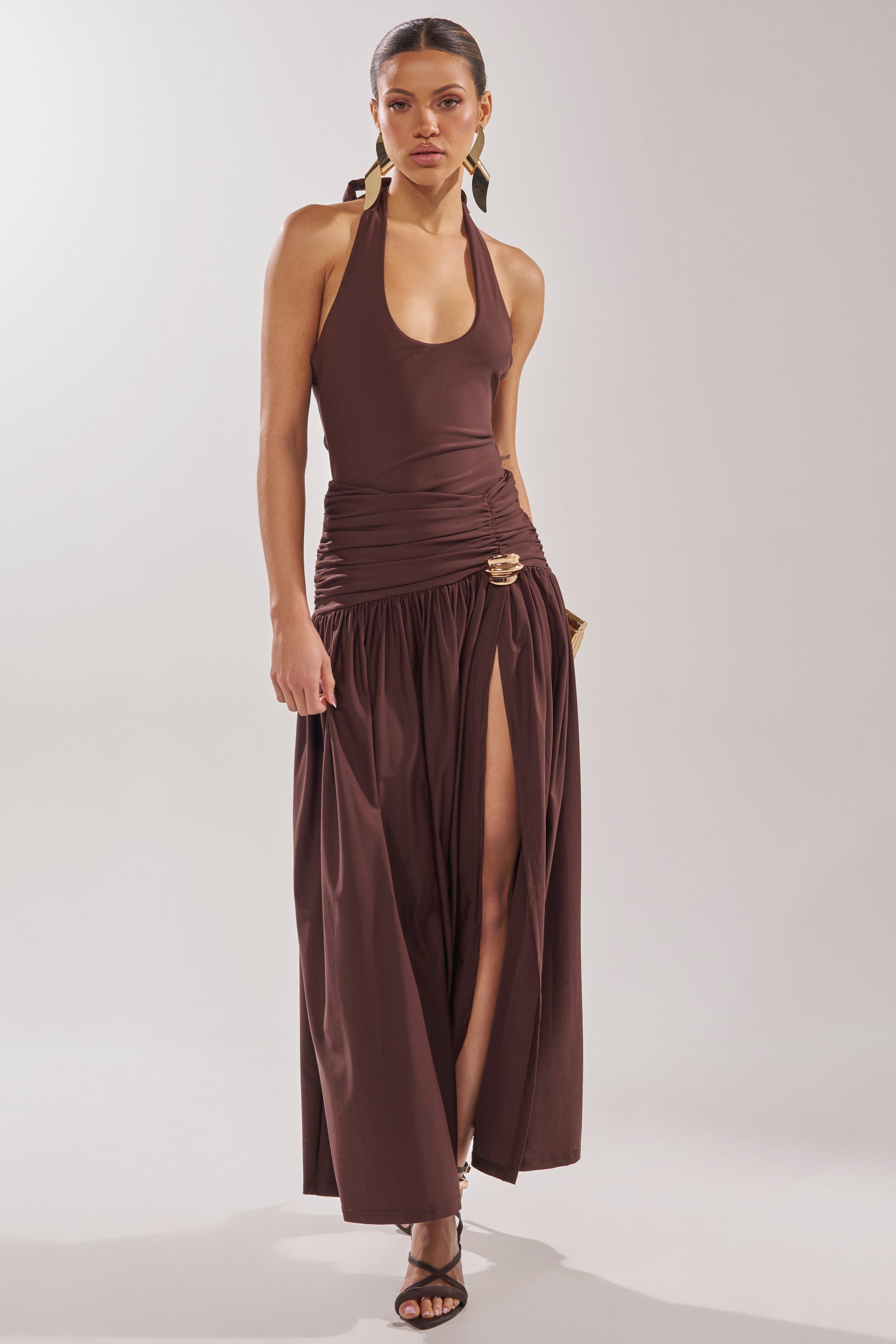 A woman wears the PERFECT RUCHED MAXI SKIRT, a brown maxi with a ruched waistband and high slit, styled with black strappy heels and gold hardware accents, standing against a plain light background. 9