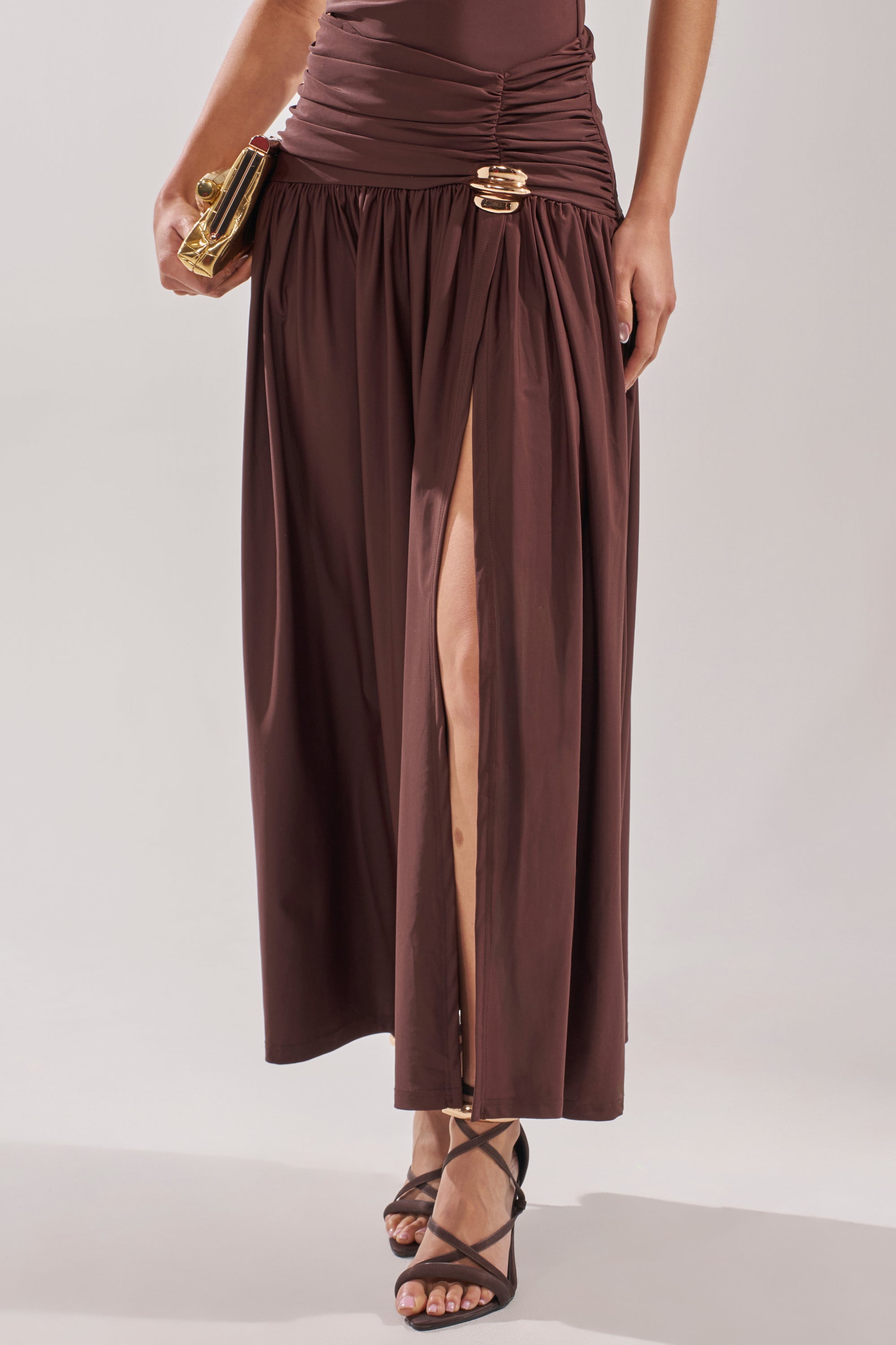 Wearing the PERFECT RUCHED MAXI SKIRT in chocolate brown with a thigh-high slit and gold hardware, paired with black strappy heels and a gold clutch. 2