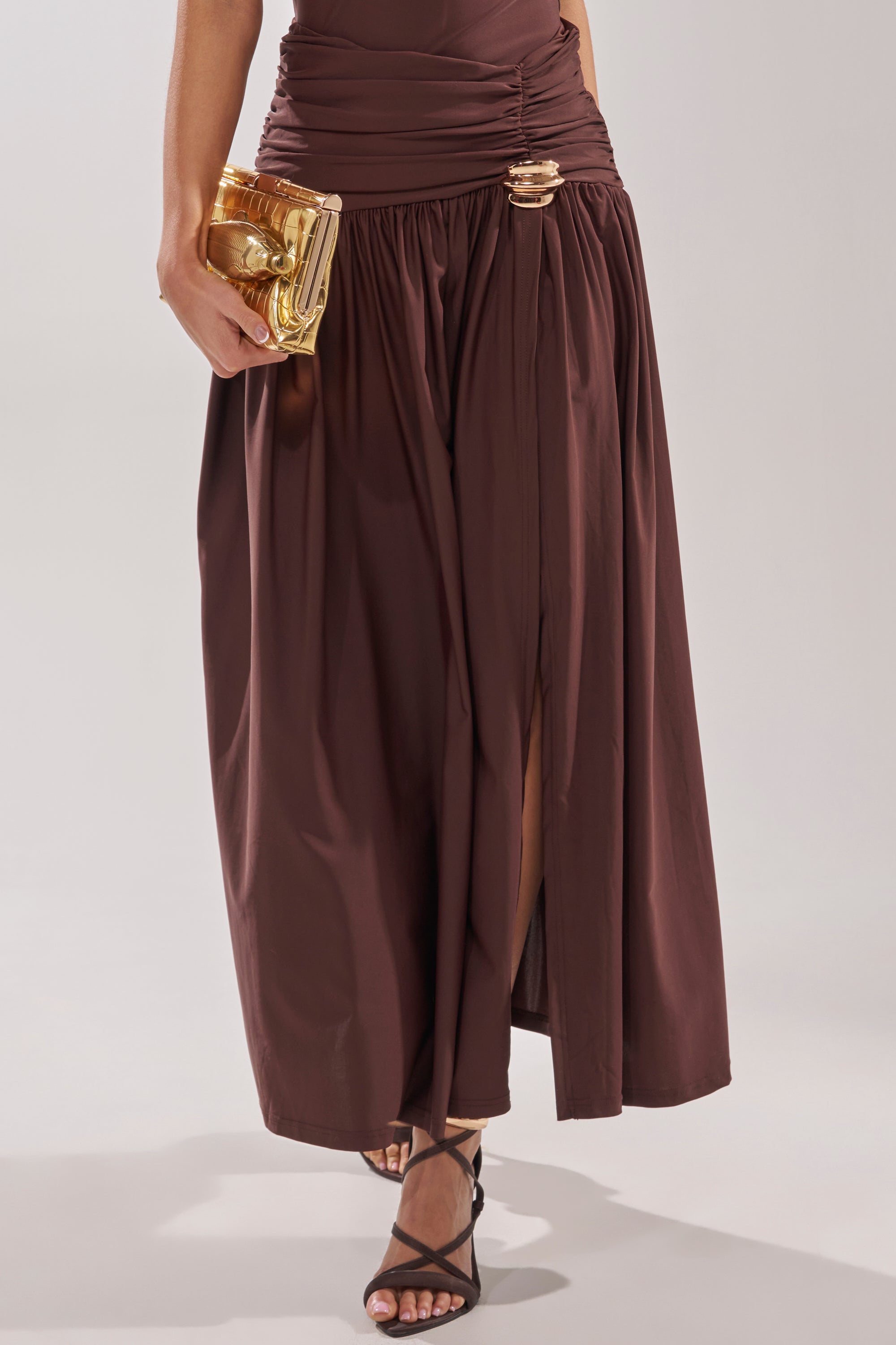 A person wearing the PERFECT RUCHED MAXI SKIRT in chocolate brown with a ruched waistband and high slit, paired with black strappy sandals and holding a shiny gold clutch. 3