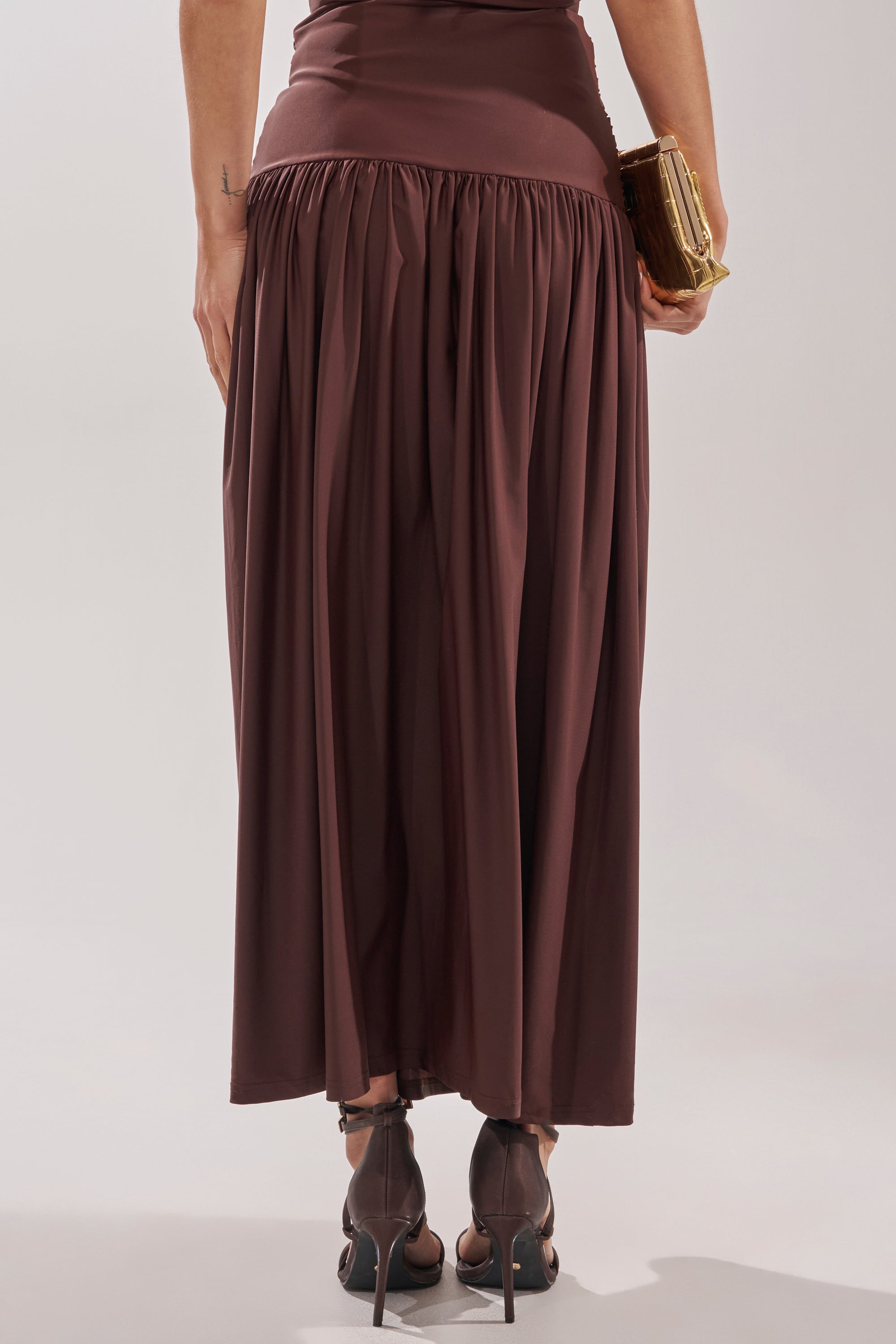 Facing away, a person wears the PERFECT RUCHED MAXI SKIRT in chocolate brown with a ruched waistband, paired with black high-heeled sandals and holding a small gold clutch featuring gold hardware against a plain light background. 5