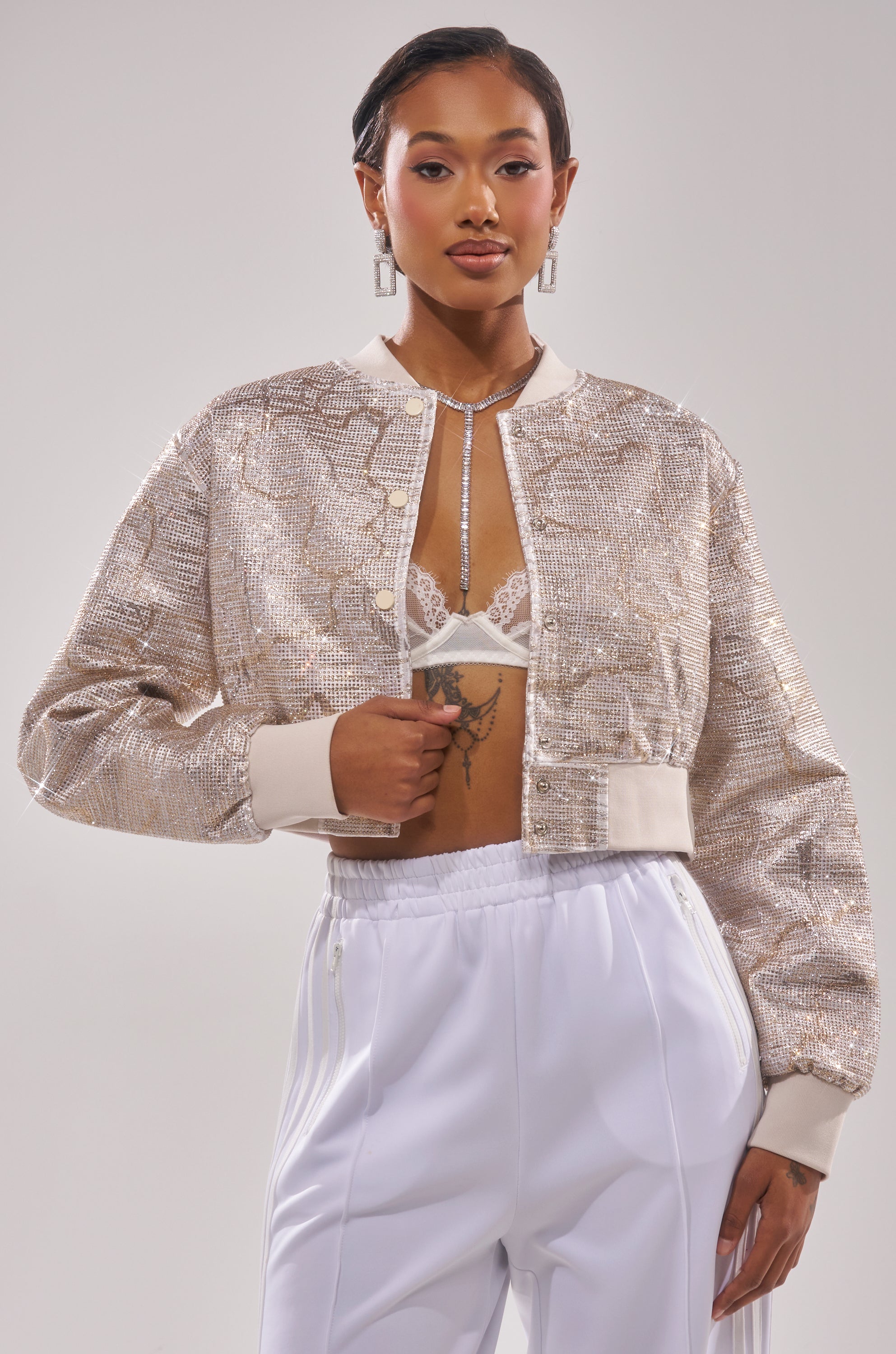 A woman poses confidently in the GOLDEN HOUR EMBELLISHED BROCADE BOMBER over a white lace bralette and high-waisted white pants, styled with geometric earrings and a statement necklace. 0