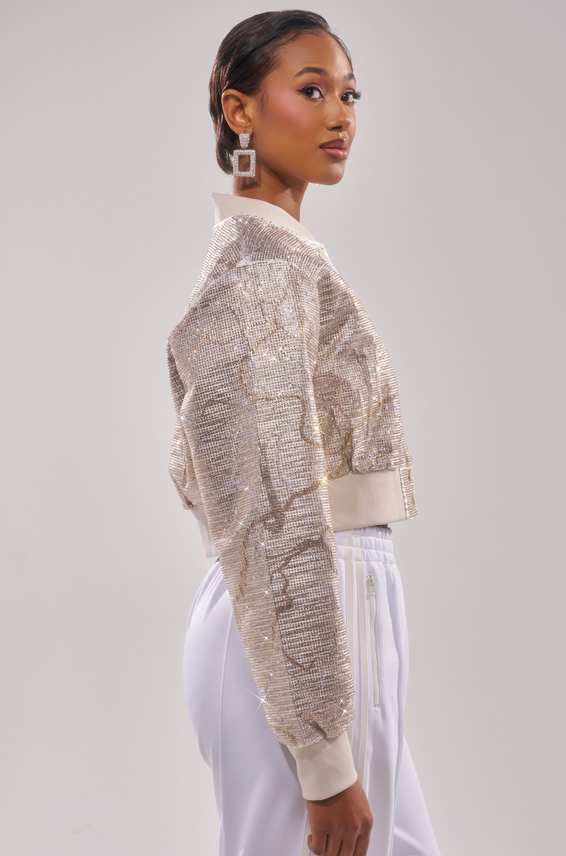 A woman in profile models the GOLDEN HOUR EMBELLISHED BROCADE BOMBER with high-waisted white pants, short slicked-back hair, geometric earrings, against a plain light background. 1