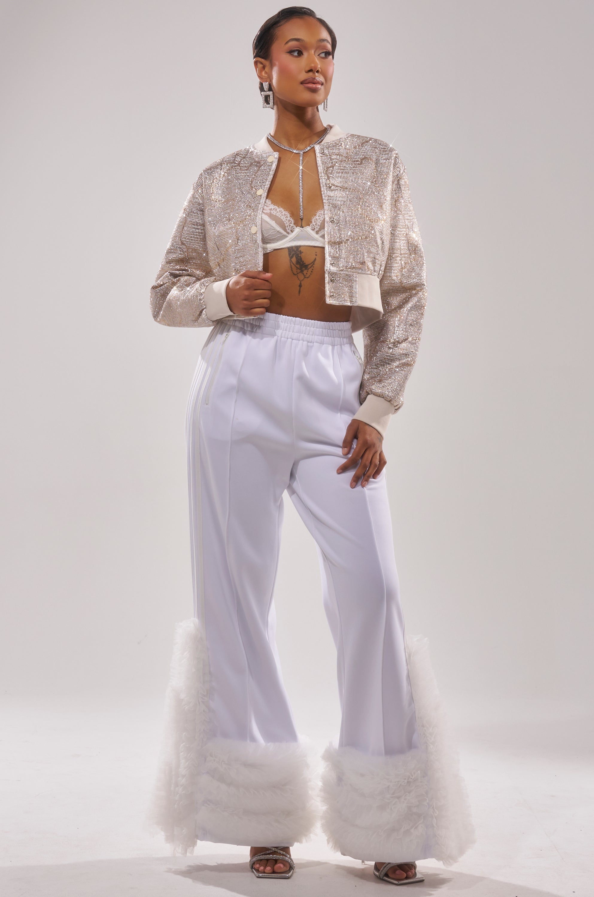 A woman poses confidently in the GOLDEN HOUR EMBELLISHED BROCADE BOMBER, a beaded bralette, and high-waisted white flared pants with fluffy hems, styled with open-toe heels against a plain light background. 3
