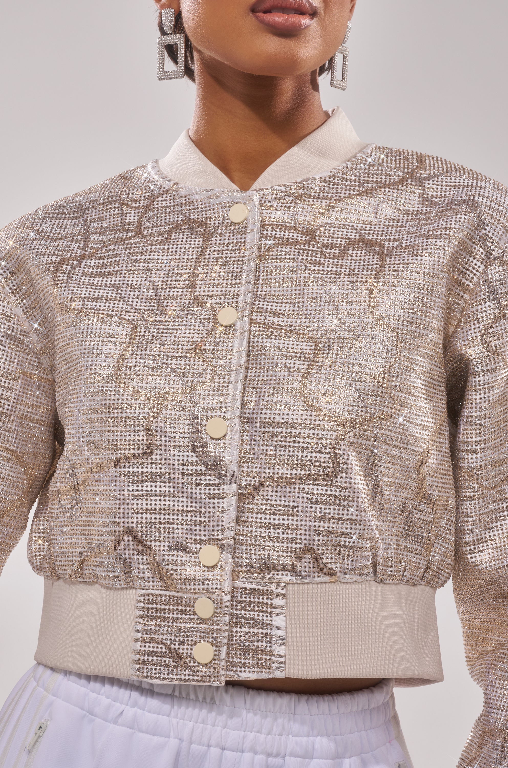 A woman wears the GOLDEN HOUR EMBELLISHED BROCADE BOMBER with a geometric pattern, gold snap buttons, cream collar and hem, styled with white pants and large square earrings. 4