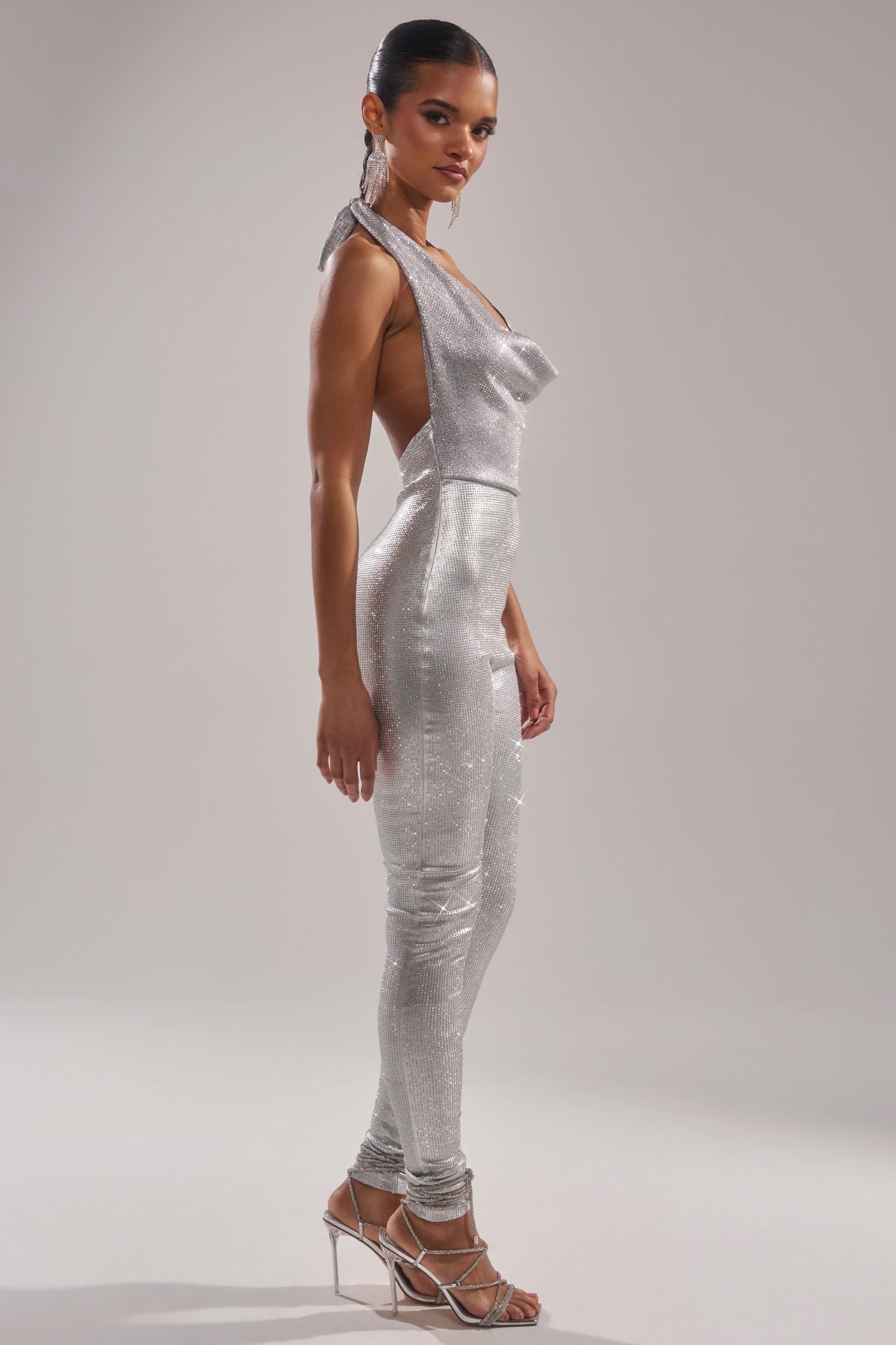 A woman poses in profile against a light background, wearing the DANCE THE NIGHT AWAY RHINESTONE JUMPSUIT with thin straps and shimmering rhinestones, paired with metallic heels. Her hair is sleek and tied back.