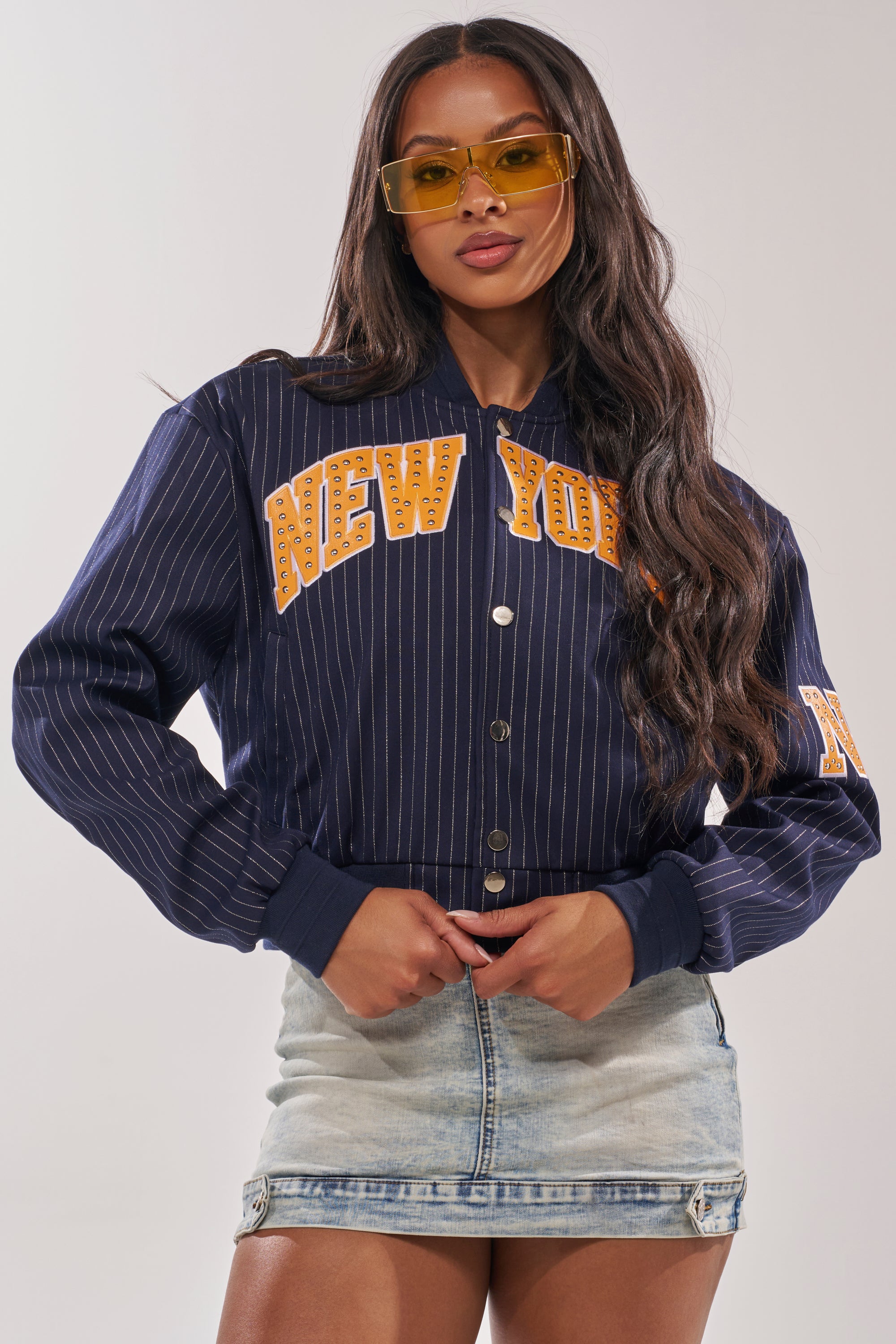 A woman in yellow-tinted glasses and a COURTSIDE PINSTRIPE BOMBER with faux leather New York appliqué pairs it with a light denim mini skirt, posing confidently against a plain background. 0