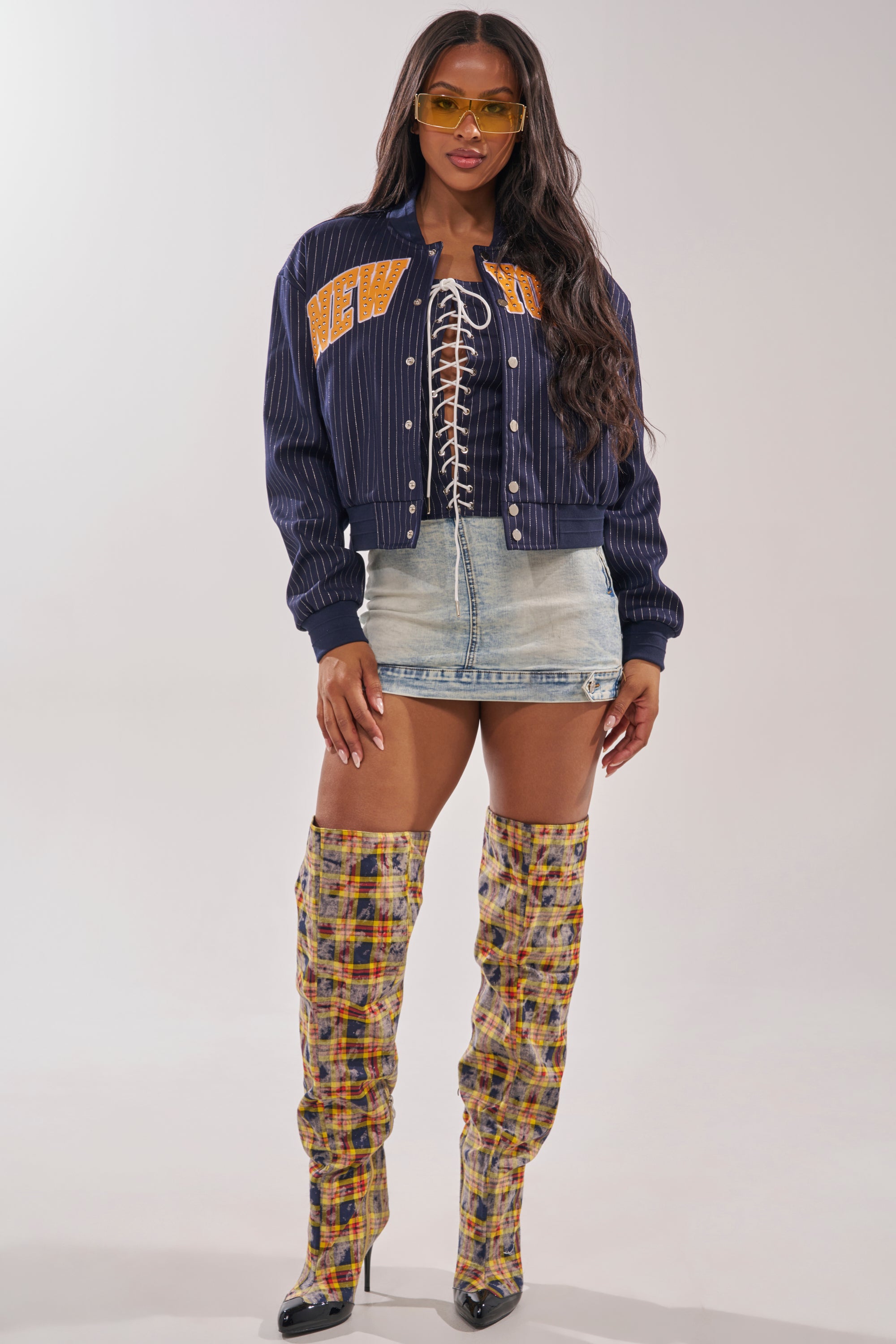 A woman wearing yellow sunglasses, a COURTSIDE PINSTRIPE BOMBER, a lace-up black top, a light blue denim mini skirt, and tall plaid over-the-knee boots stands facing forward against a plain background. 9