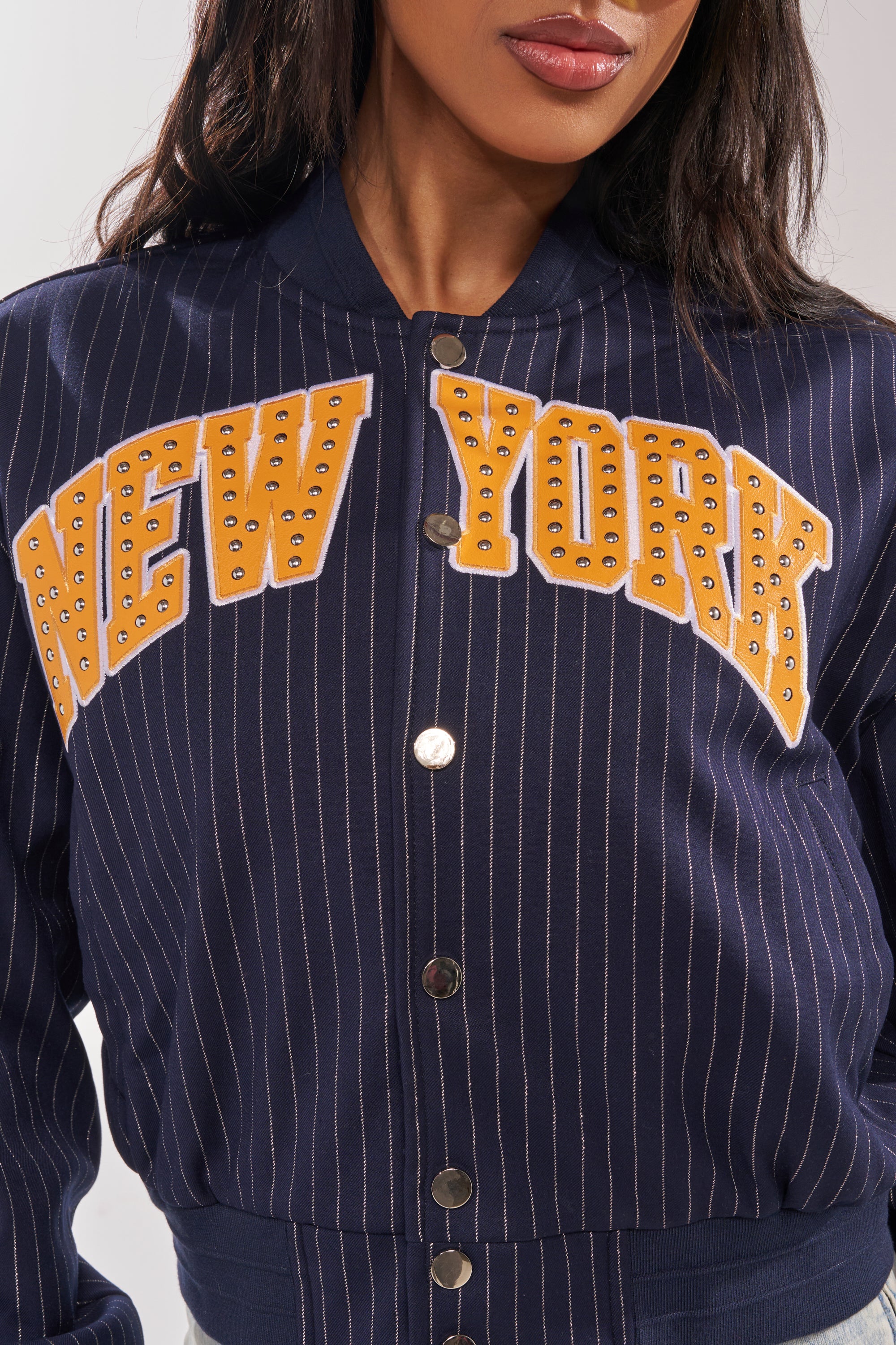 A woman wears the COURTSIDE PINSTRIPE BOMBER, a navy blue pinstripe jacket with bold yellow studded "NEW YORK" lettering and metallic snap buttons. Only her torso and lower face are shown. 11