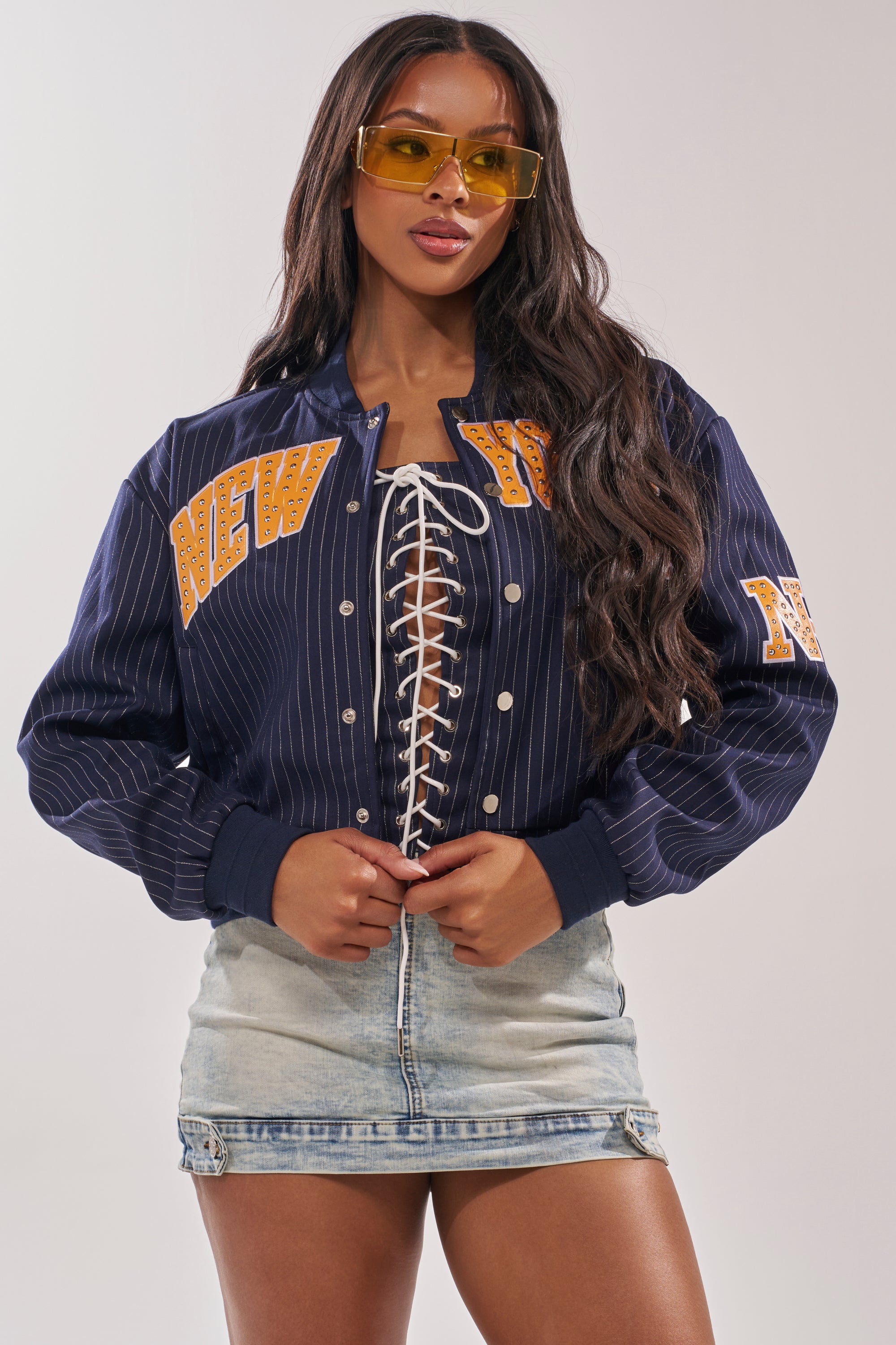 A woman wearing yellow-tinted sunglasses, the COURTSIDE PINSTRIPE BOMBER with a faux leather New York appliqué and lace-up front, and a light denim mini skirt stands against a plain background. 1