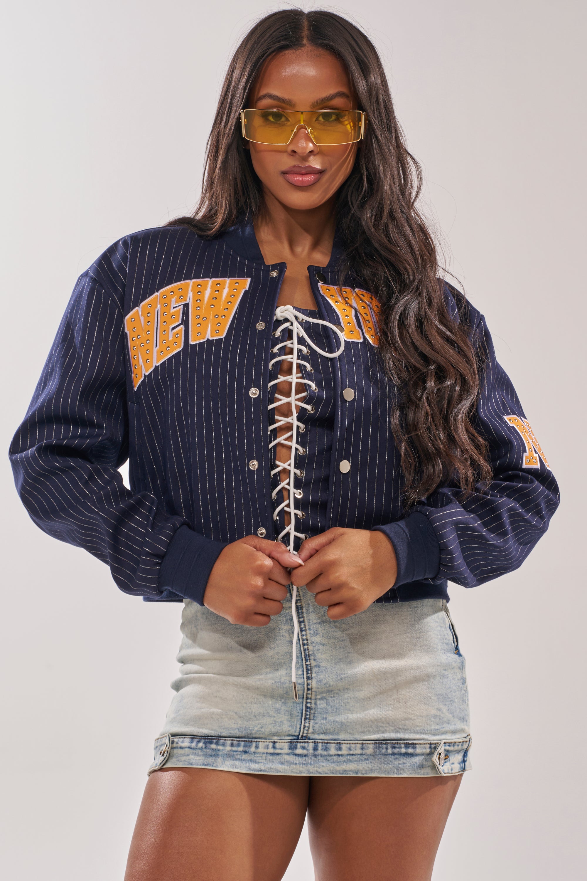 A woman in yellow sunglasses and a light denim mini skirt poses, holding the COURTSIDE PINSTRIPE BOMBER, featuring faux leather New York appliqué. 2
