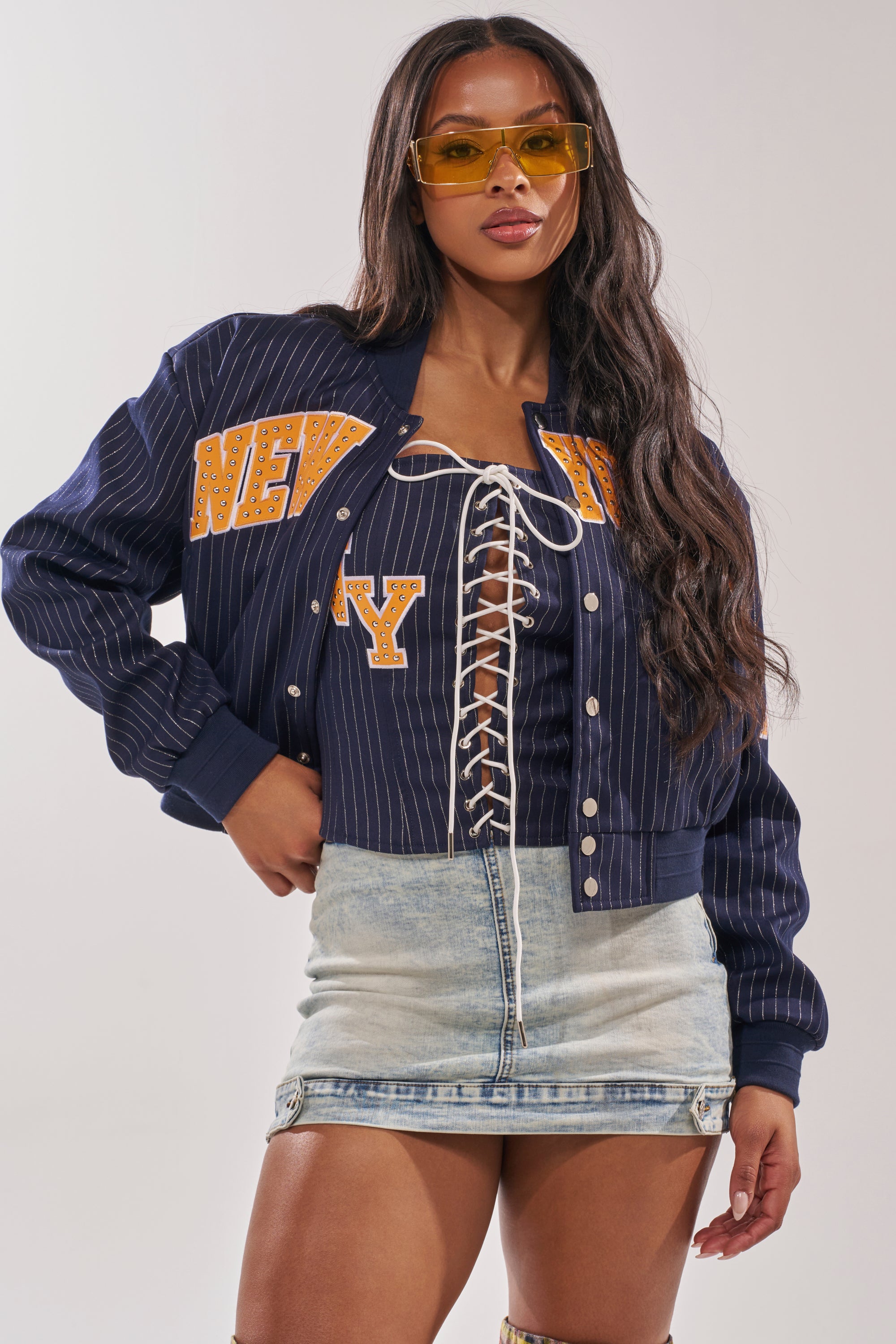 A woman in yellow sunglasses, a COURTSIDE PINSTRIPE BOMBER with "NEW YORK" lettering, a lace-up white top, and a light blue denim mini skirt stands with one hand on her hip. 3