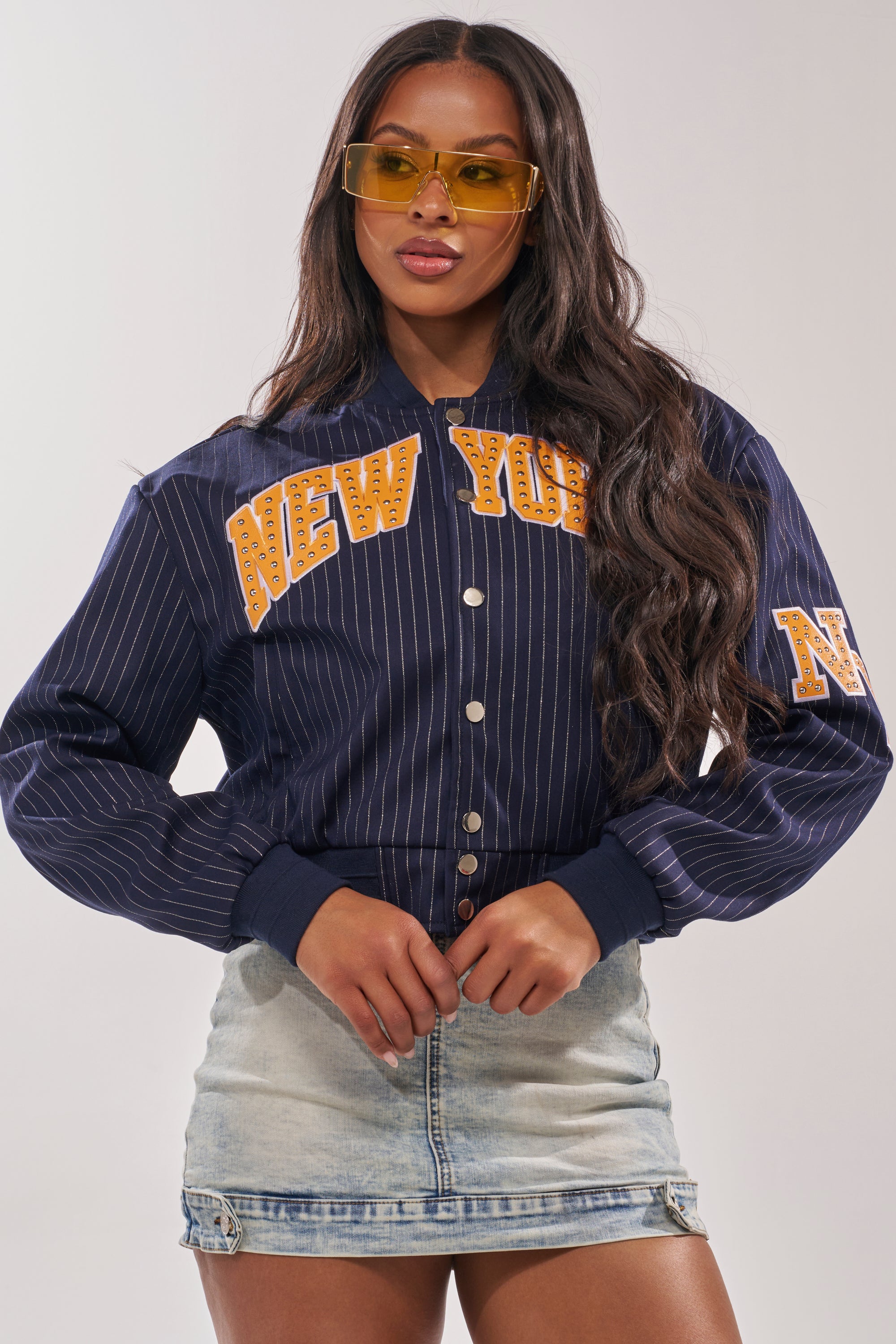A woman wearing yellow-tinted sunglasses, a faux leather COURTSIDE PINSTRIPE BOMBER, and a light denim mini skirt stands against a plain background. 4