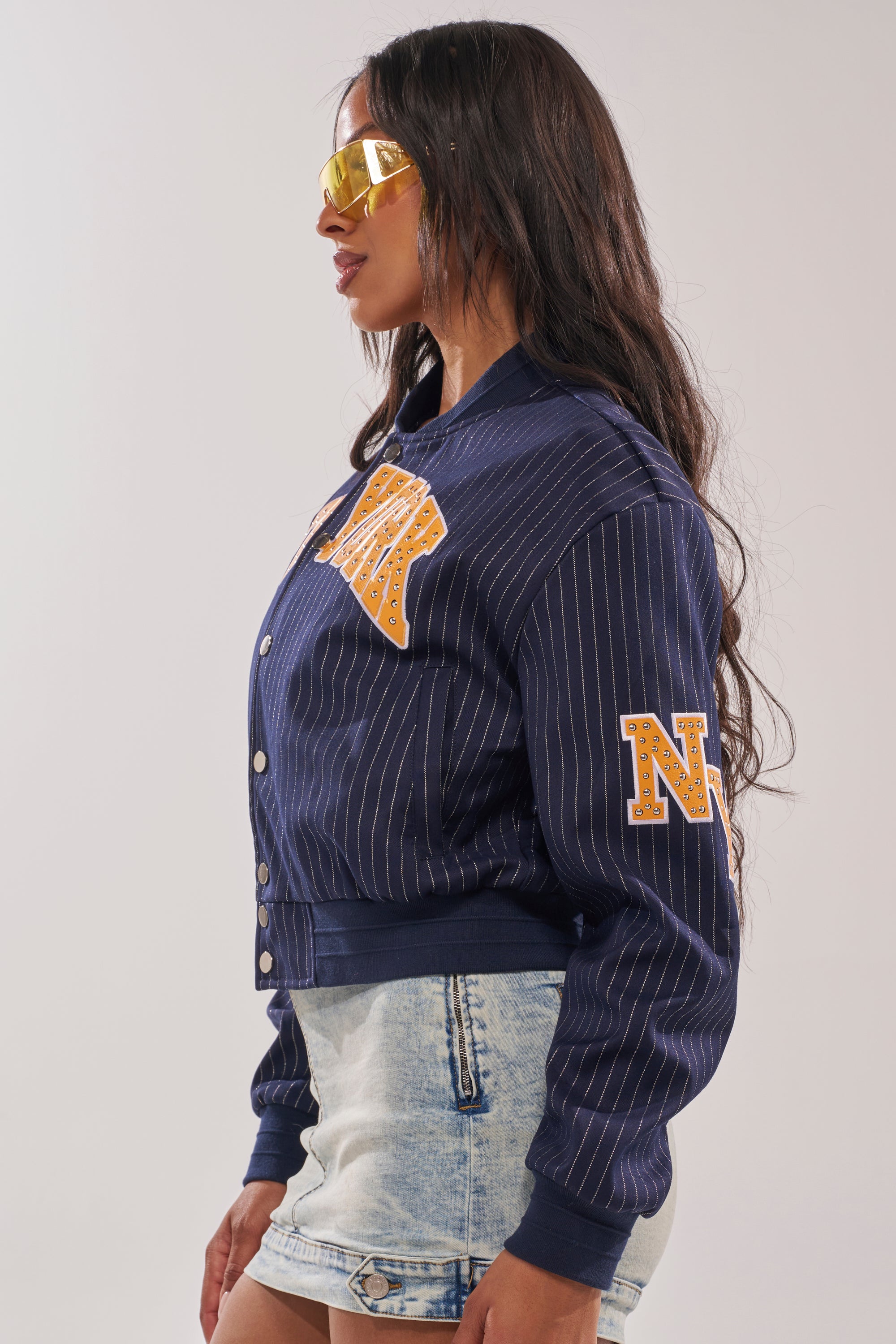 A woman in yellow sunglasses and the COURTSIDE PINSTRIPE BOMBER with bold NEW YORK appliqué pairs it with light blue denim shorts, standing in profile against a plain background for a sporty street style look. 6