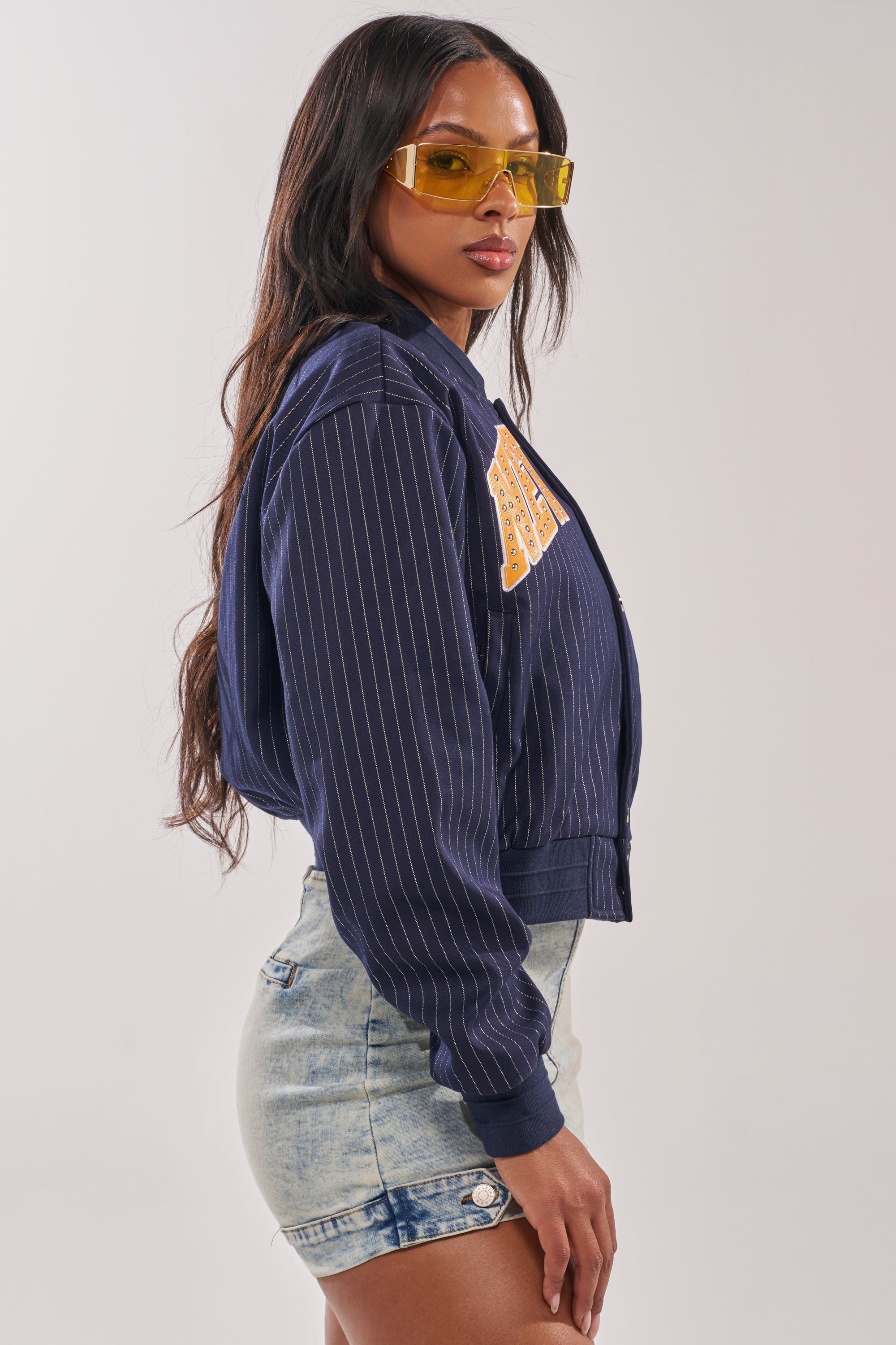 Woman stands sideways in a COURTSIDE PINSTRIPE BOMBER with a NEW YORK patch, sporting yellow-tinted sunglasses and light denim shorts. Her long, wavy hair and neutral expression evoke sporty street style against a plain background. 7