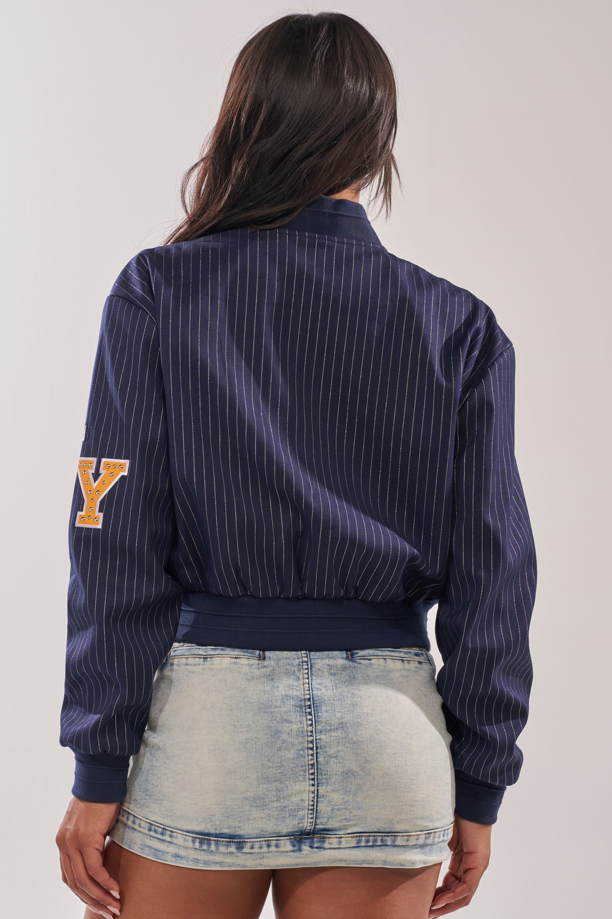 A woman faces away, wearing the COURTSIDE PINSTRIPE BOMBER in navy with a yellow "Y" patch on the sleeve and a light-wash distressed denim mini skirt. 8