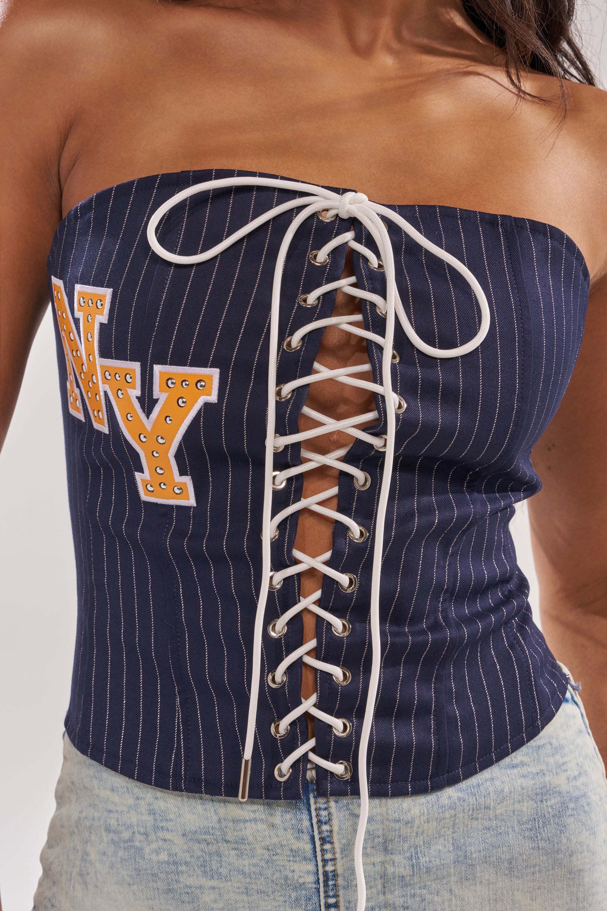A woman channels city-girl style in the COURTSIDE LACE UP CORSET—a navy pinstripe, strapless top with white lace-up front and bold yellow "NY" lettering—perfectly paired with light-wash denim jeans. 9