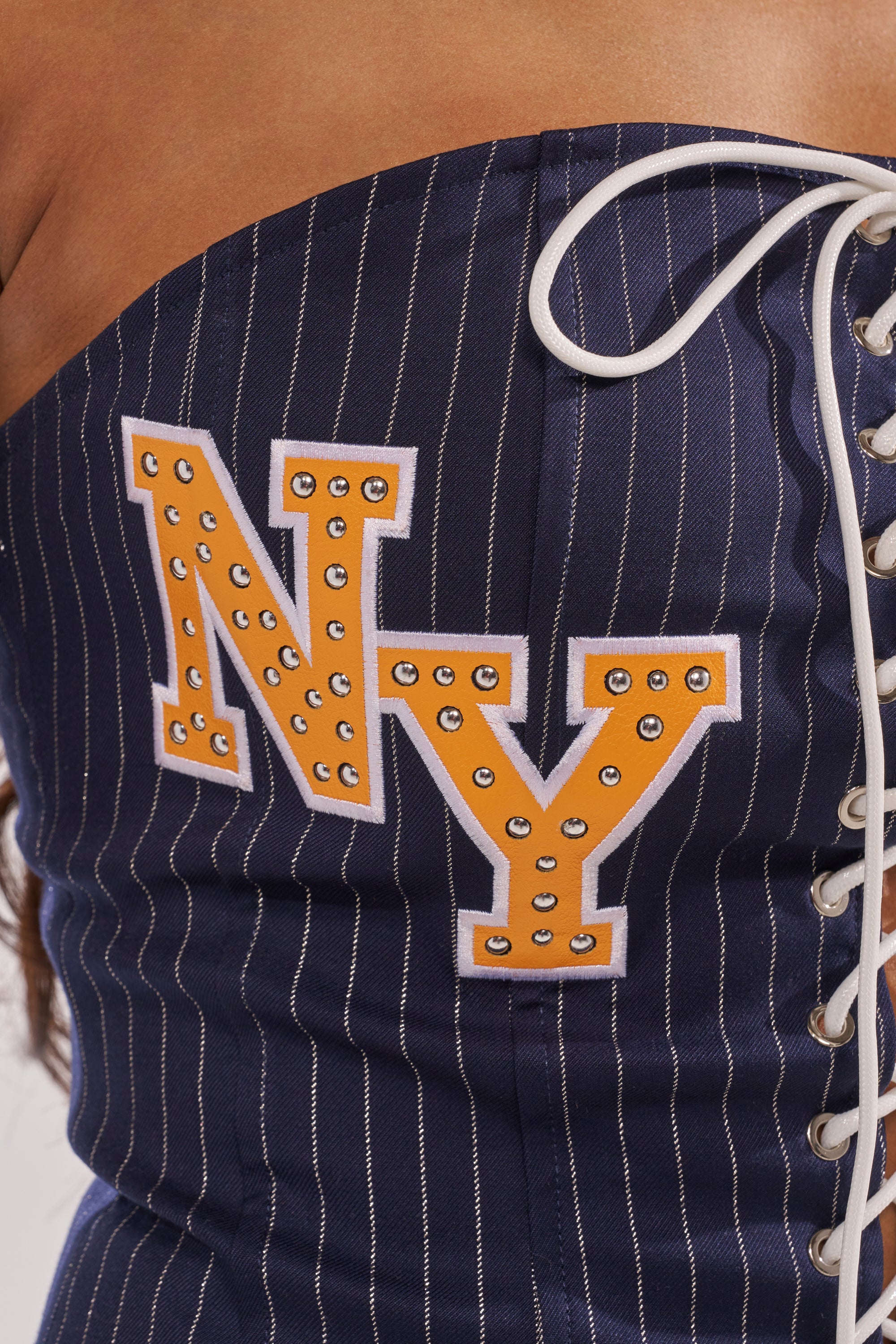 Close-up of the COURTSIDE LACE UP CORSET: navy pinstripe, strapless neckline, white laces, and orange "NY" with silver studs on the side—ideal for city-girl style. 10