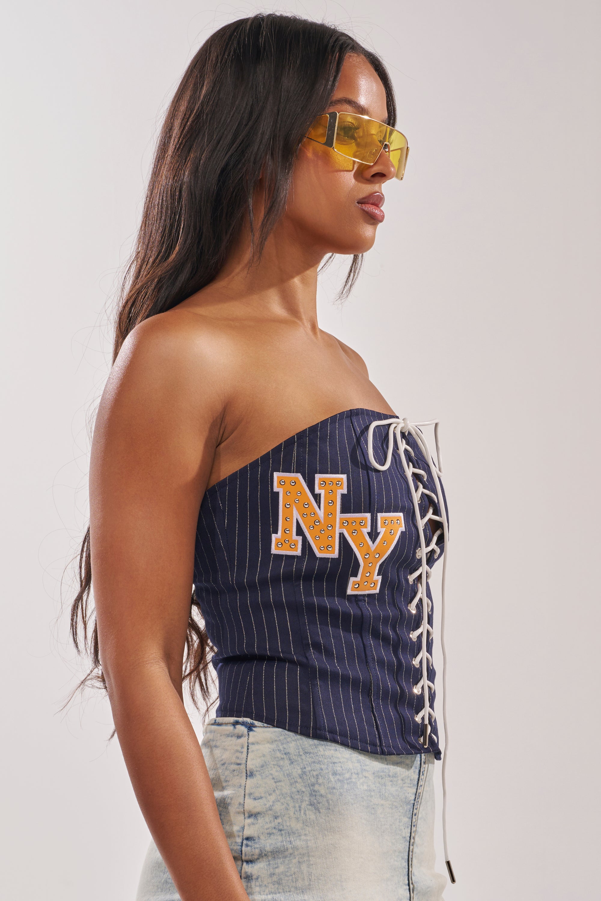 A woman with long dark hair channels city-girl style in yellow sunglasses, the COURTSIDE LACE UP CORSET with "NY" lettering and lace-up details, and light blue jeans against a pale background. 4