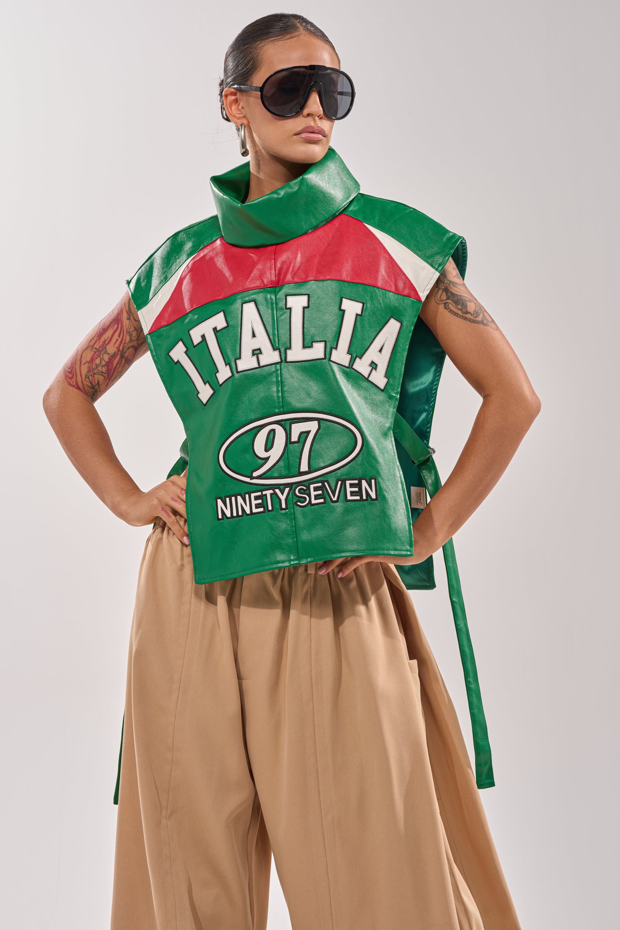 A woman stands confidently with one hand on her hip, showcasing bold streetwear layering in the CATCHING FLIGHTS EMBROIDERED VEST—an oversized green, red, and white sleeveless statement piece styled with wide-leg beige pants and sunglasses. 0