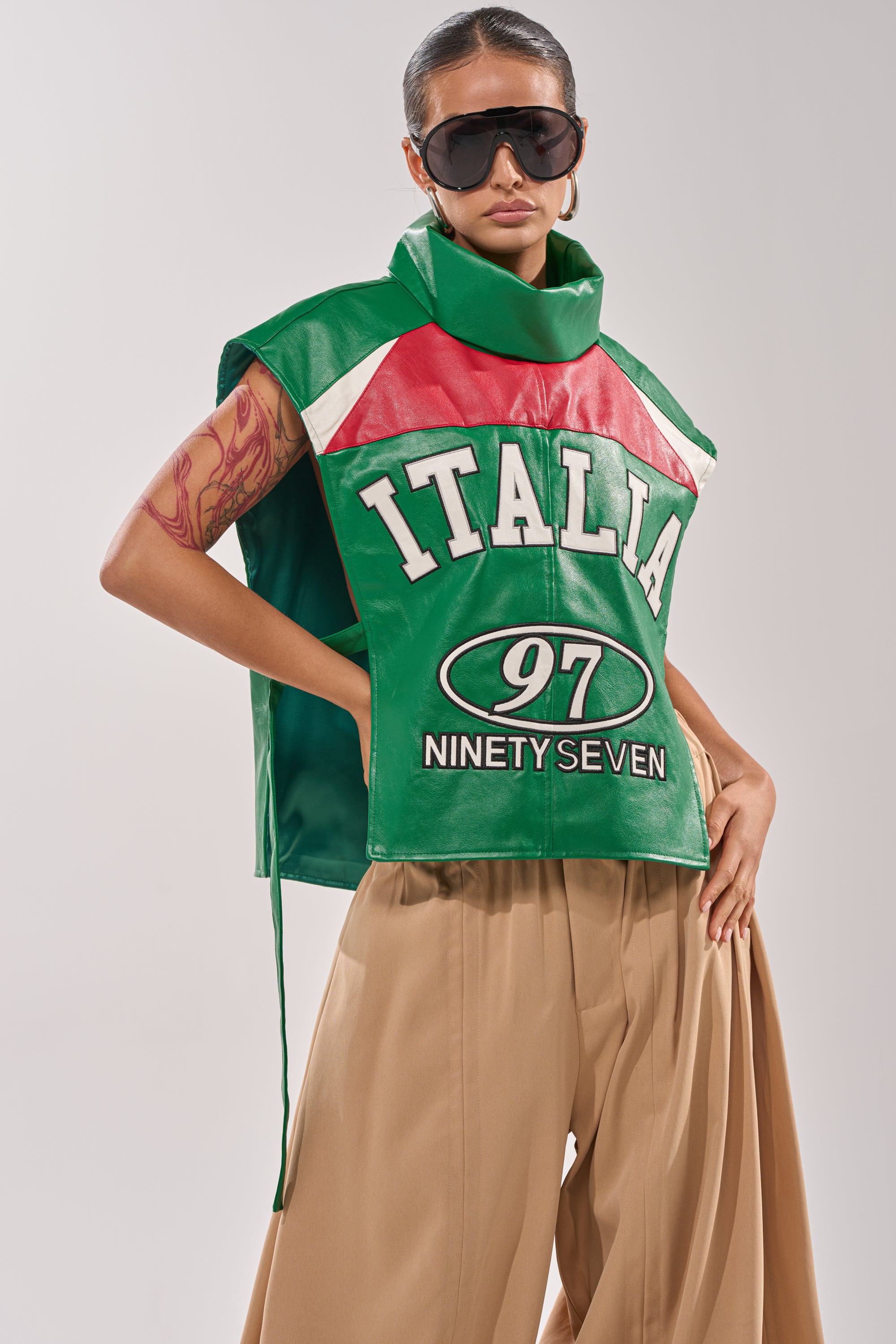 A person wears oversized sunglasses, wide tan pants, and the CATCHING FLIGHTS EMBROIDERED VEST in green, red, and white with "ITALIA 97 NINETY SEVEN" text, posing with one arm bent to reveal a forearm tattoo. 1