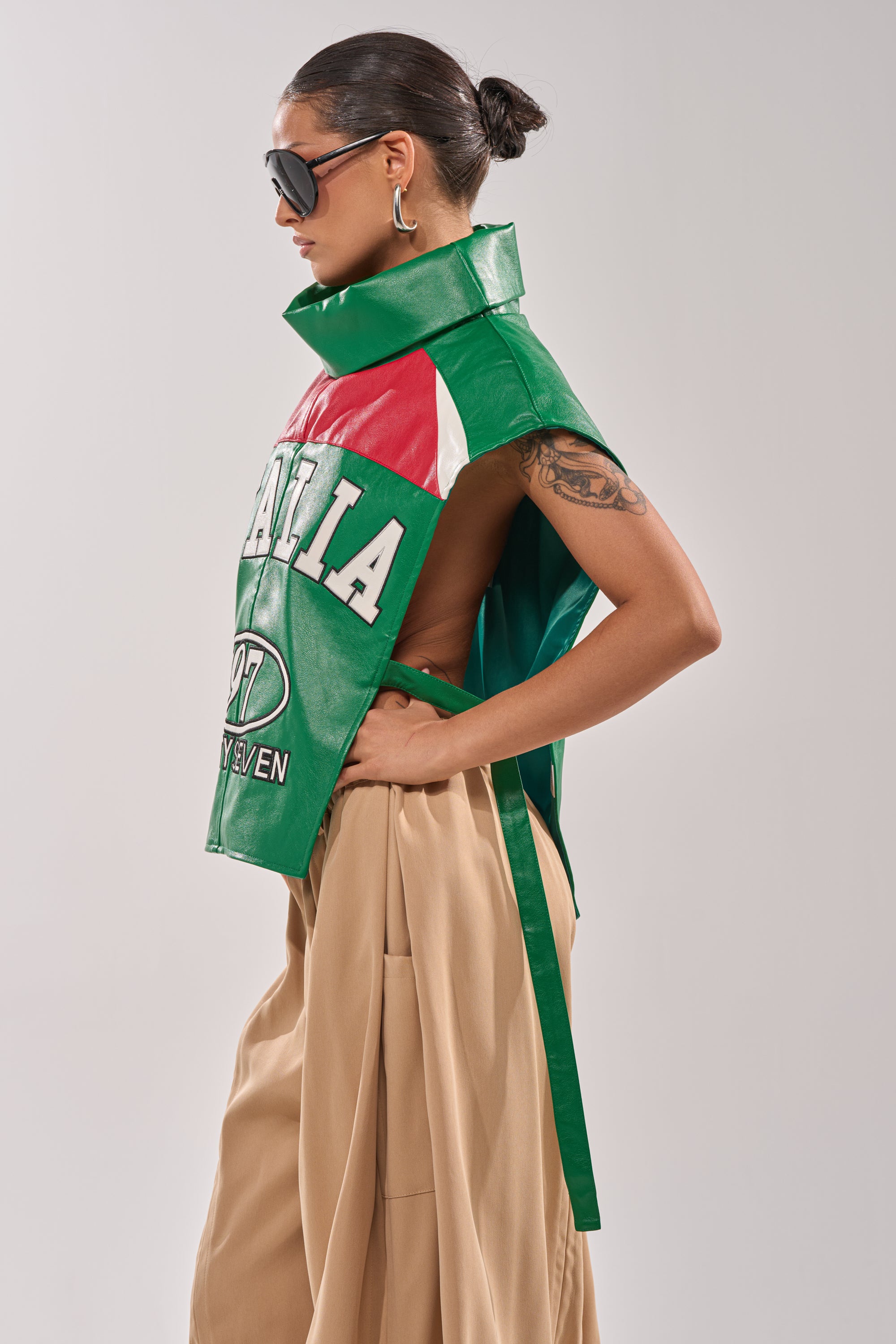 A woman in beige pleated pants and a bun hairstyle models the CATCHING FLIGHTS EMBROIDERED VEST—green, red, and white with "ITALIA" and "07"—styled with hoop earrings and large sunglasses against a plain background. 2