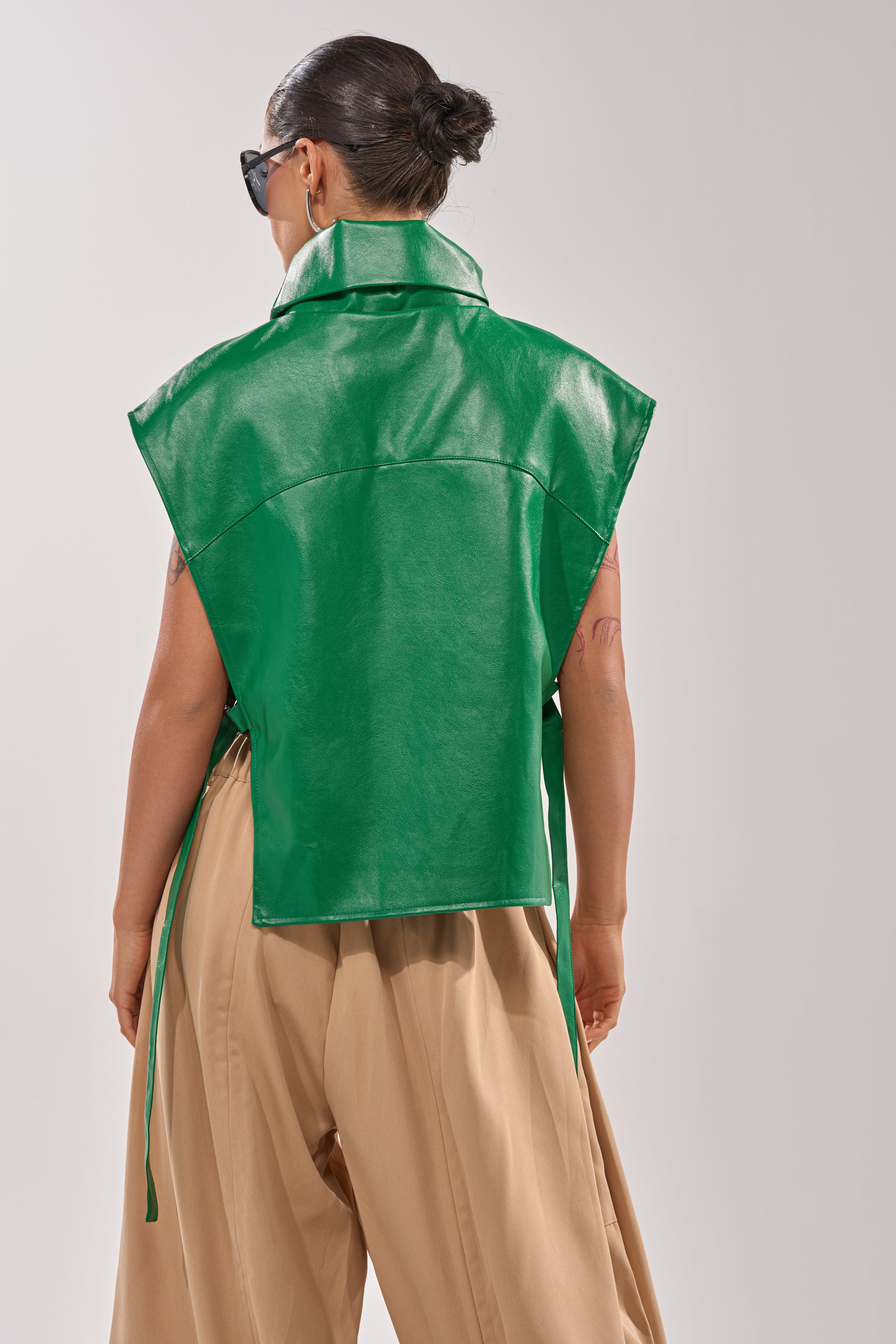 A person with dark hair in a bun, wearing sunglasses and the CATCHING FLIGHTS EMBROIDERED VEST—a sleeveless green faux leather vest with side ties—paired with wide-legged beige pants, is seen from behind against a plain background. 3