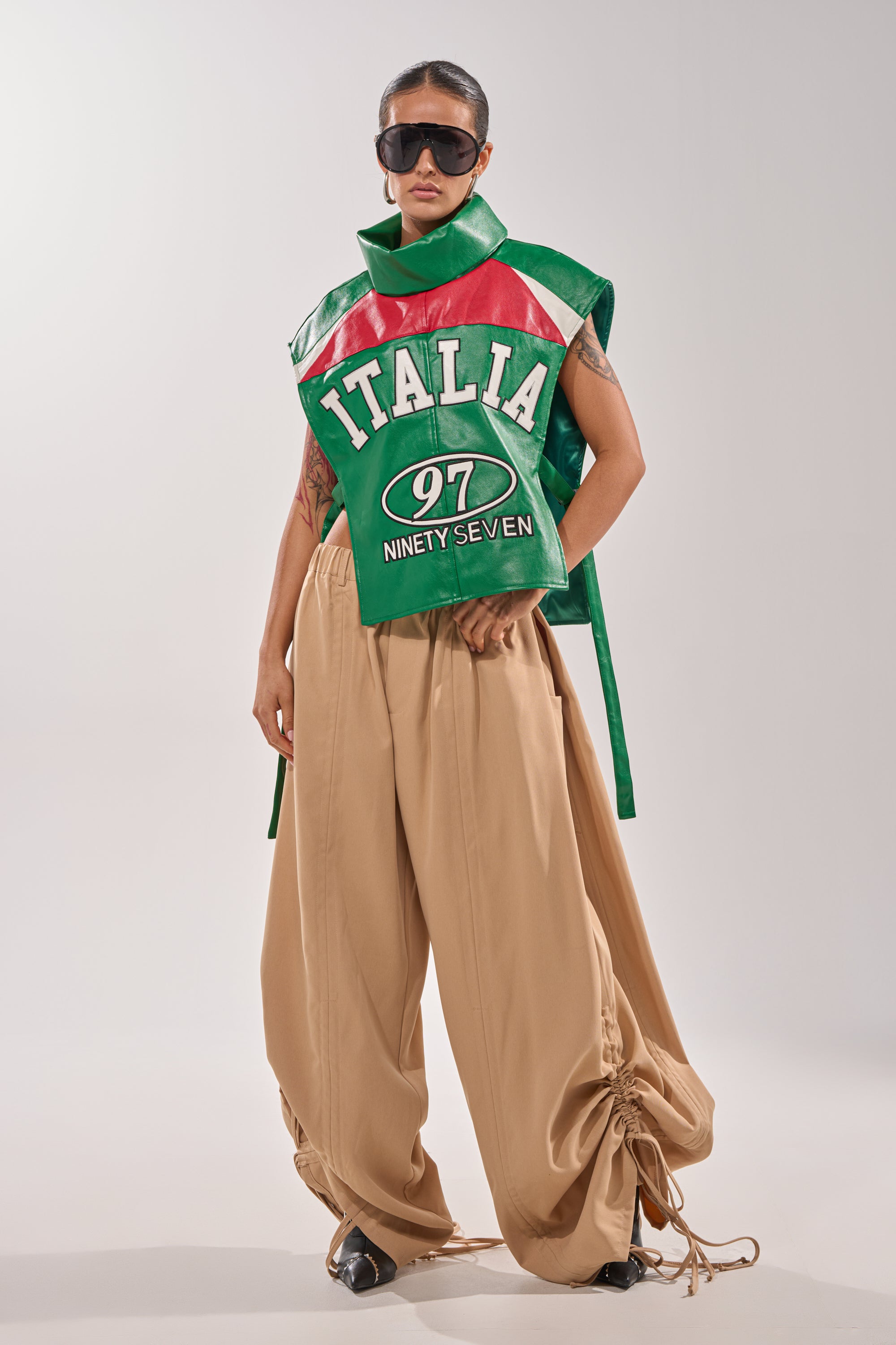 A person wearing oversized tan pants with gathered hems, the CATCHING FLIGHTS EMBROIDERED VEST, and large dark sunglasses stands against a plain background. 6