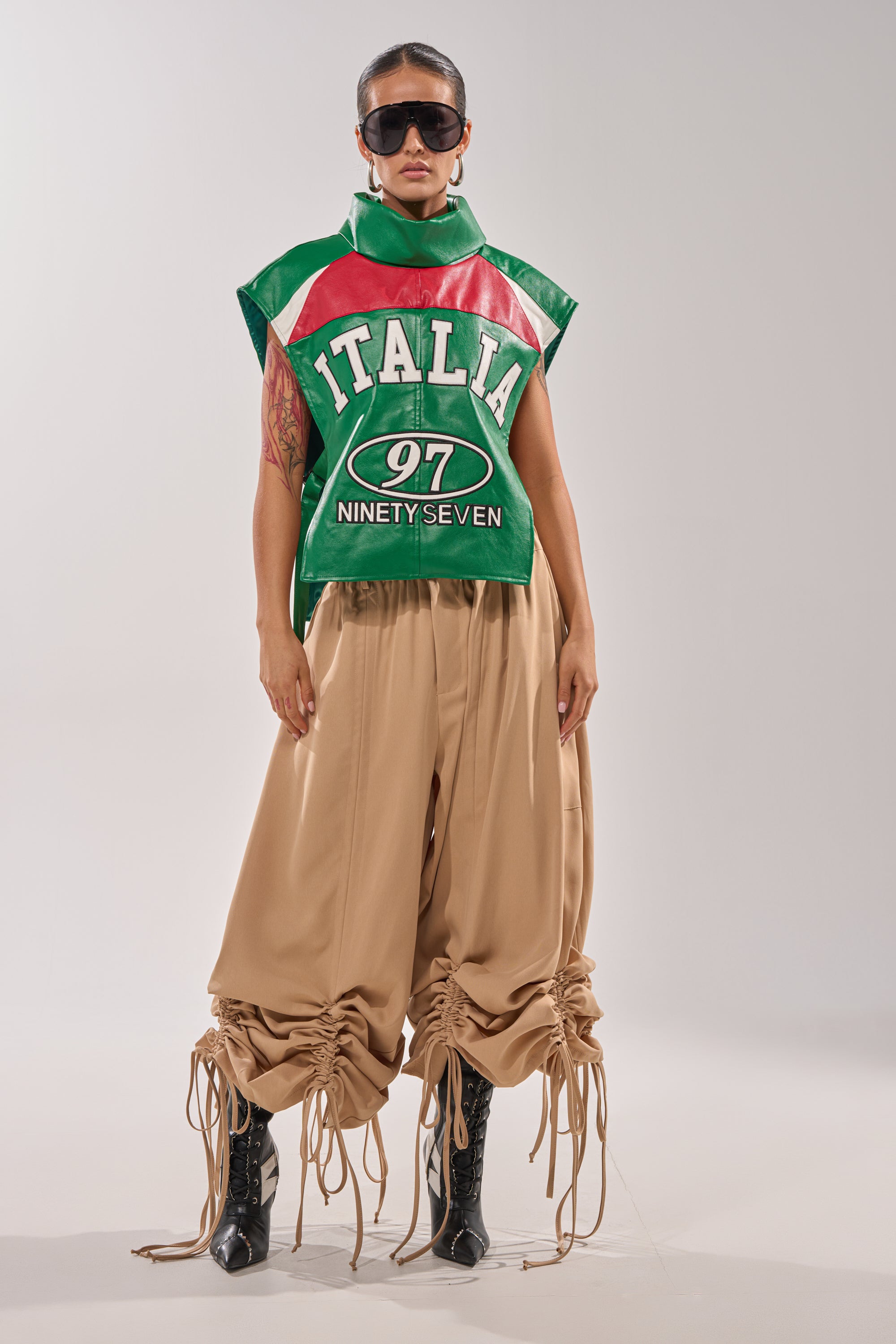 A person wears oversized sunglasses, wide beige drawstring pants, black pointed boots, and the CATCHING FLIGHTS EMBROIDERED VEST—a green, red, and white sleeveless layer with bold "ITALIA 97 NINETY SEVEN" text—for a striking streetwear look. 7