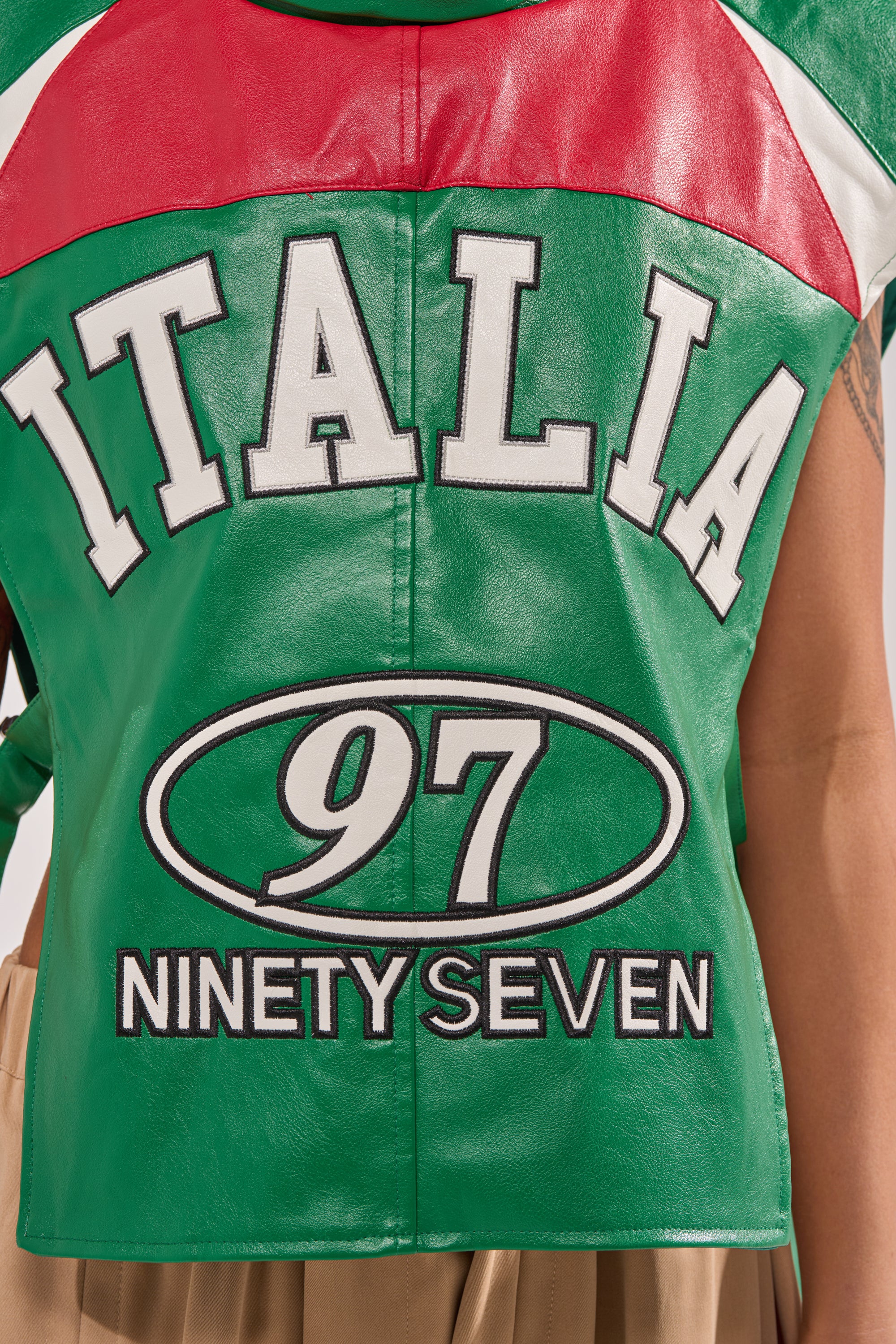 A person stands with their back to the camera, showcasing the CATCHING FLIGHTS EMBROIDERED VEST—a green, red, and white sleeveless statement piece with "ITALIA" and "97 NINETY SEVEN" boldly displayed on the back for standout streetwear style. 8