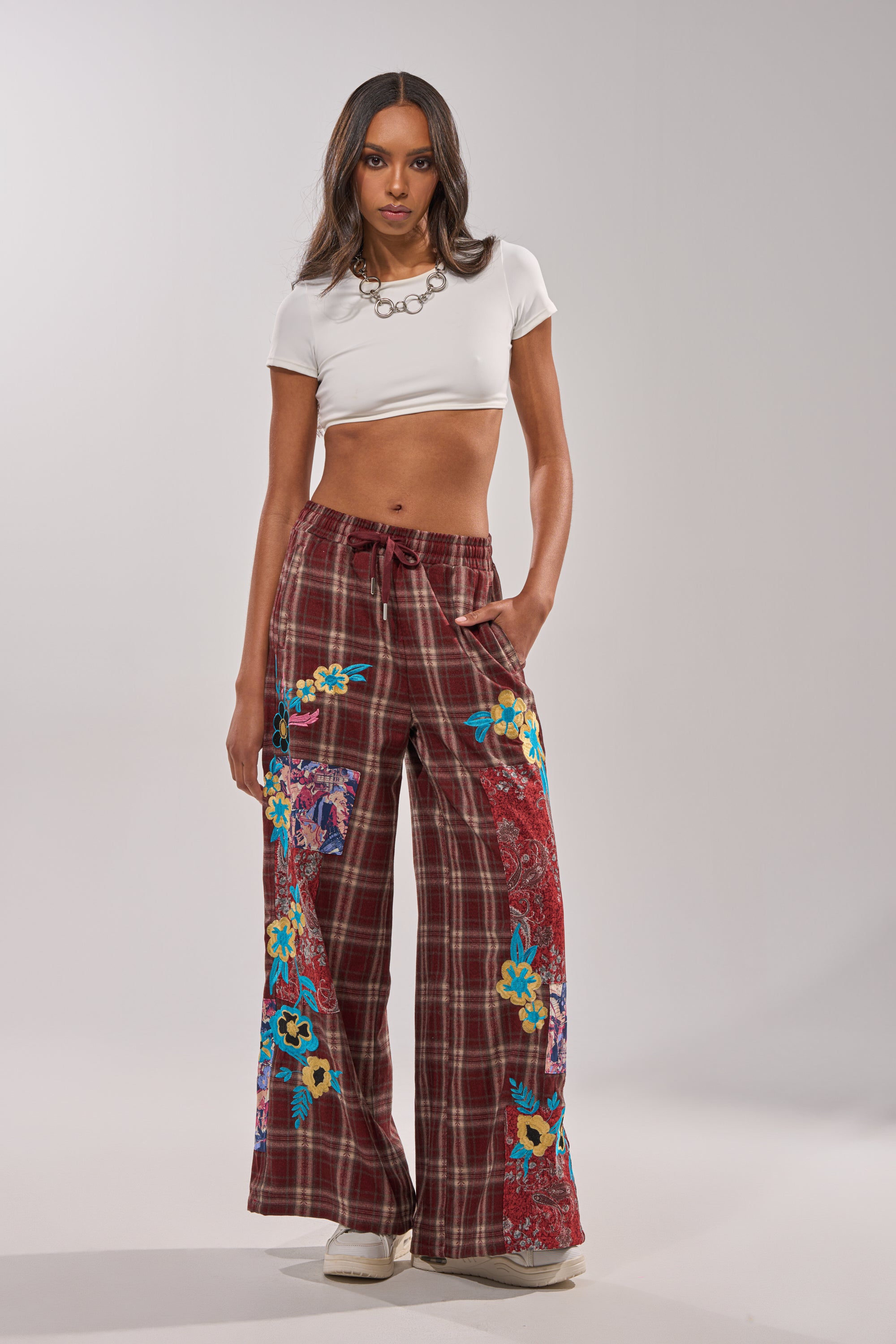 A woman with long hair and a neutral expression wears the KRIYSTA FLORAL PATCH PLAID PANT—wide-leg maroon plaid pants with colorful floral patchwork—paired with a white cropped t-shirt, white shoes, and a chunky chain necklace against a plain background. 0