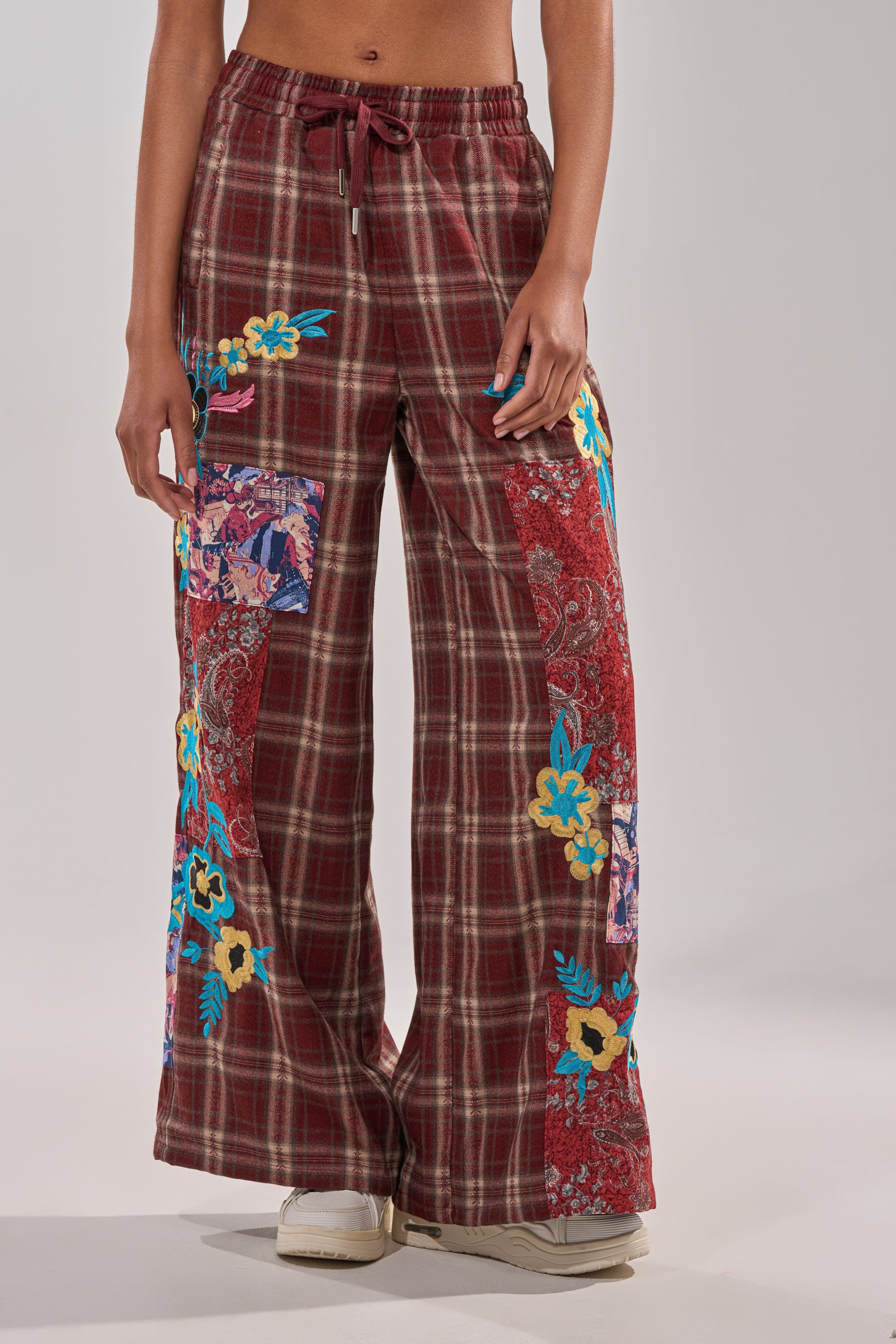 A person stands against a plain background wearing KRIYSTA FLORAL PATCH PLAID PANT—wide-leg, high-waisted pants with colorful floral and plaid patches, a drawstring waist, and paired with white sneakers. 1