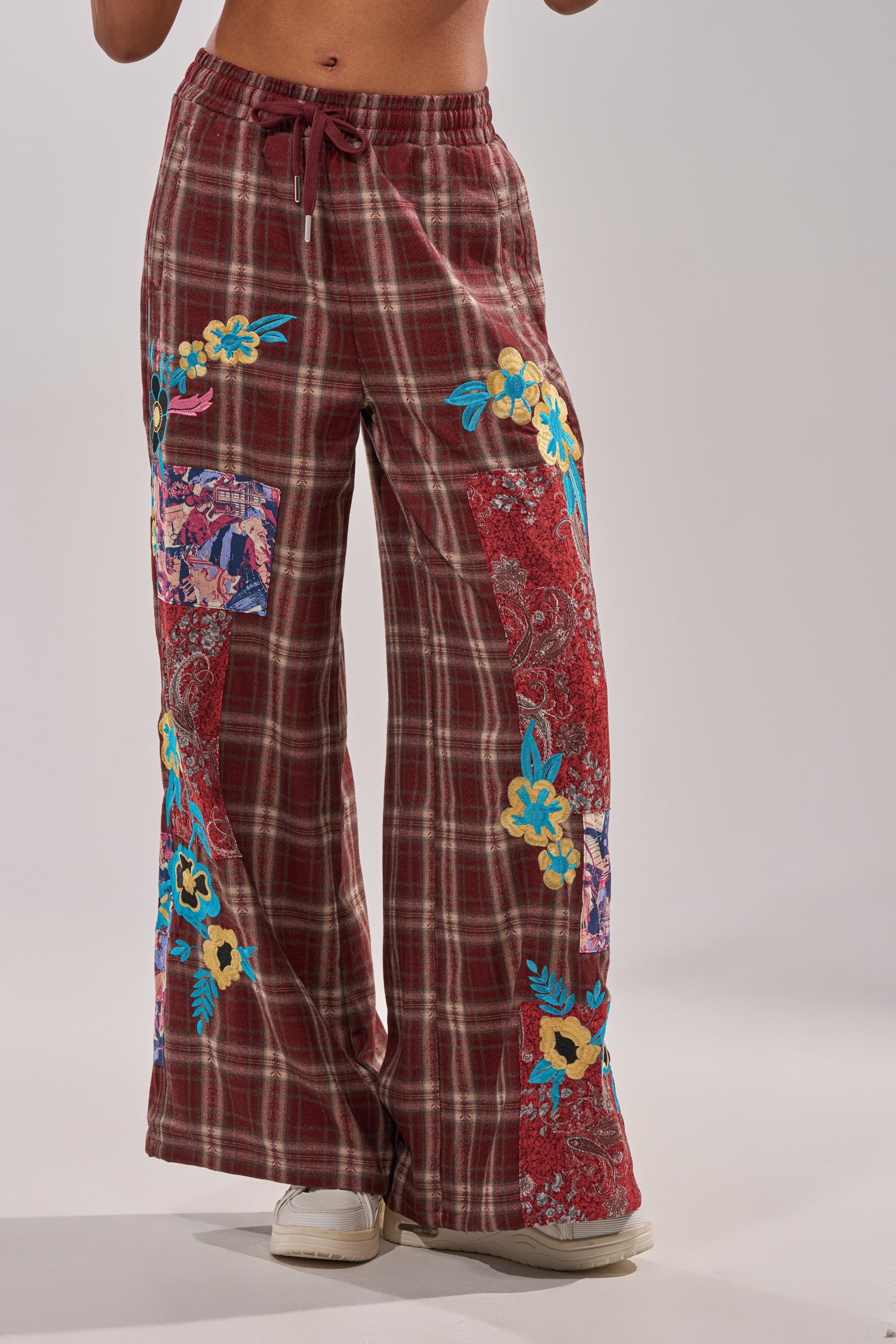 Only the lower torso and legs are visible, showcasing the KRIYSTA FLORAL PATCH PLAID PANT—wide leg, high waisted pants with plaid patchwork and colorful floral and paisley embroidery, set against a light background. 2