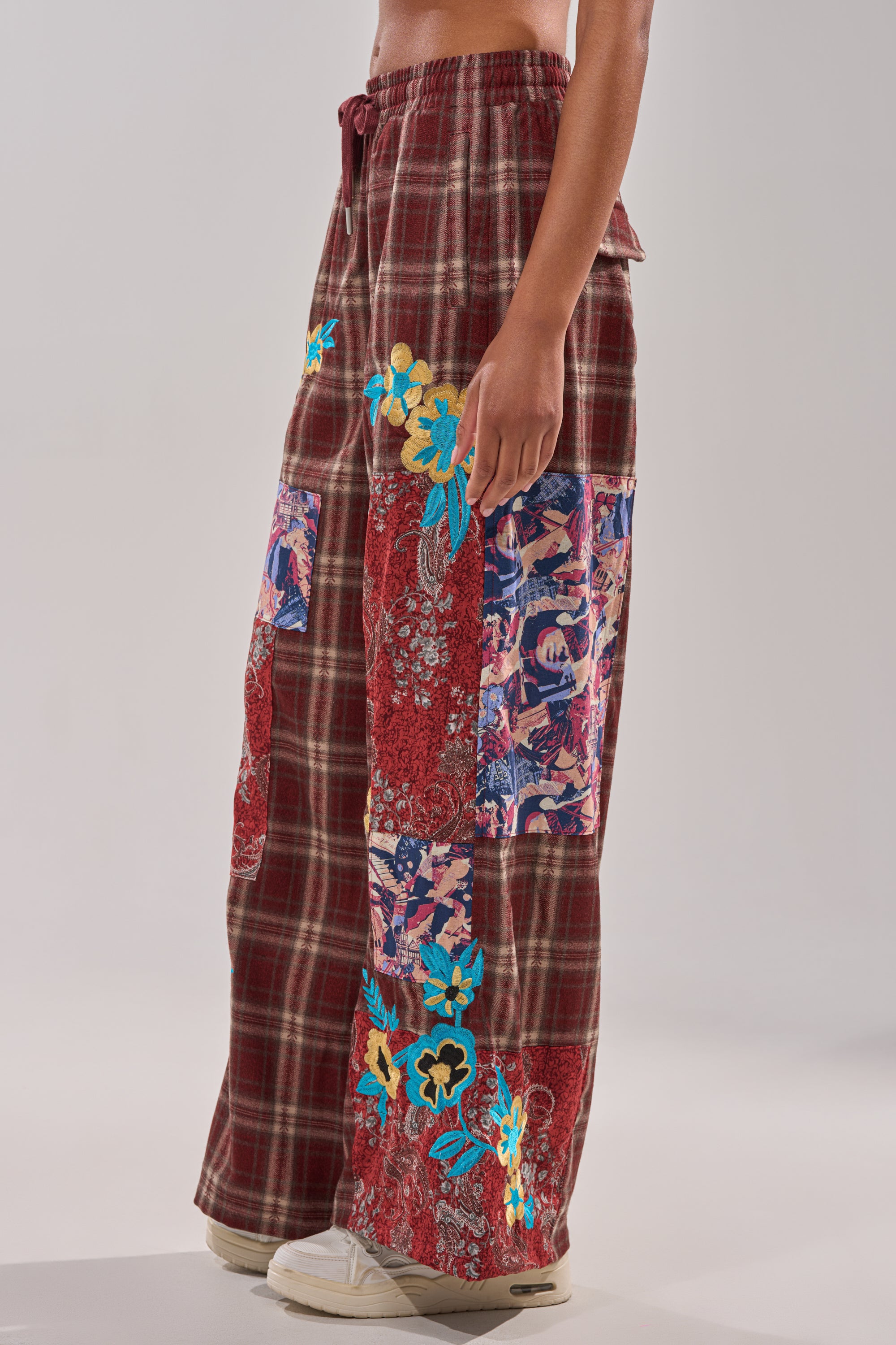 A person models the KRIYSTA FLORAL PATCH PLAID PANT, featuring wide-leg plaid with a red base and vibrant floral patches, styled with white sneakers against a neutral background. 3