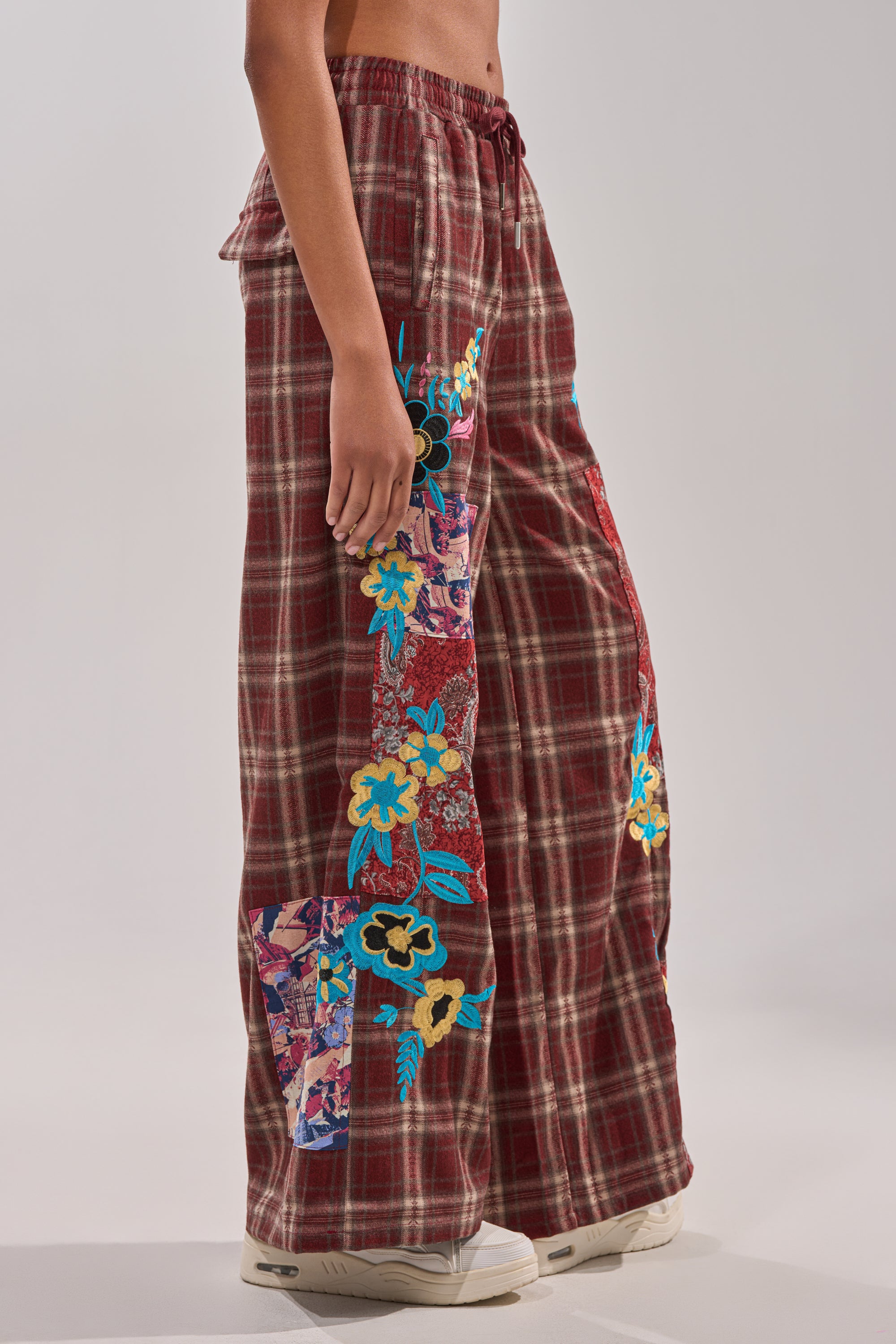 Wearing the KRIYSTA FLORAL PATCH PLAID PANT—loose, wide-leg plaid pants with colorful floral patchwork and embroidery—a person in white sneakers stands against a plain background. 4