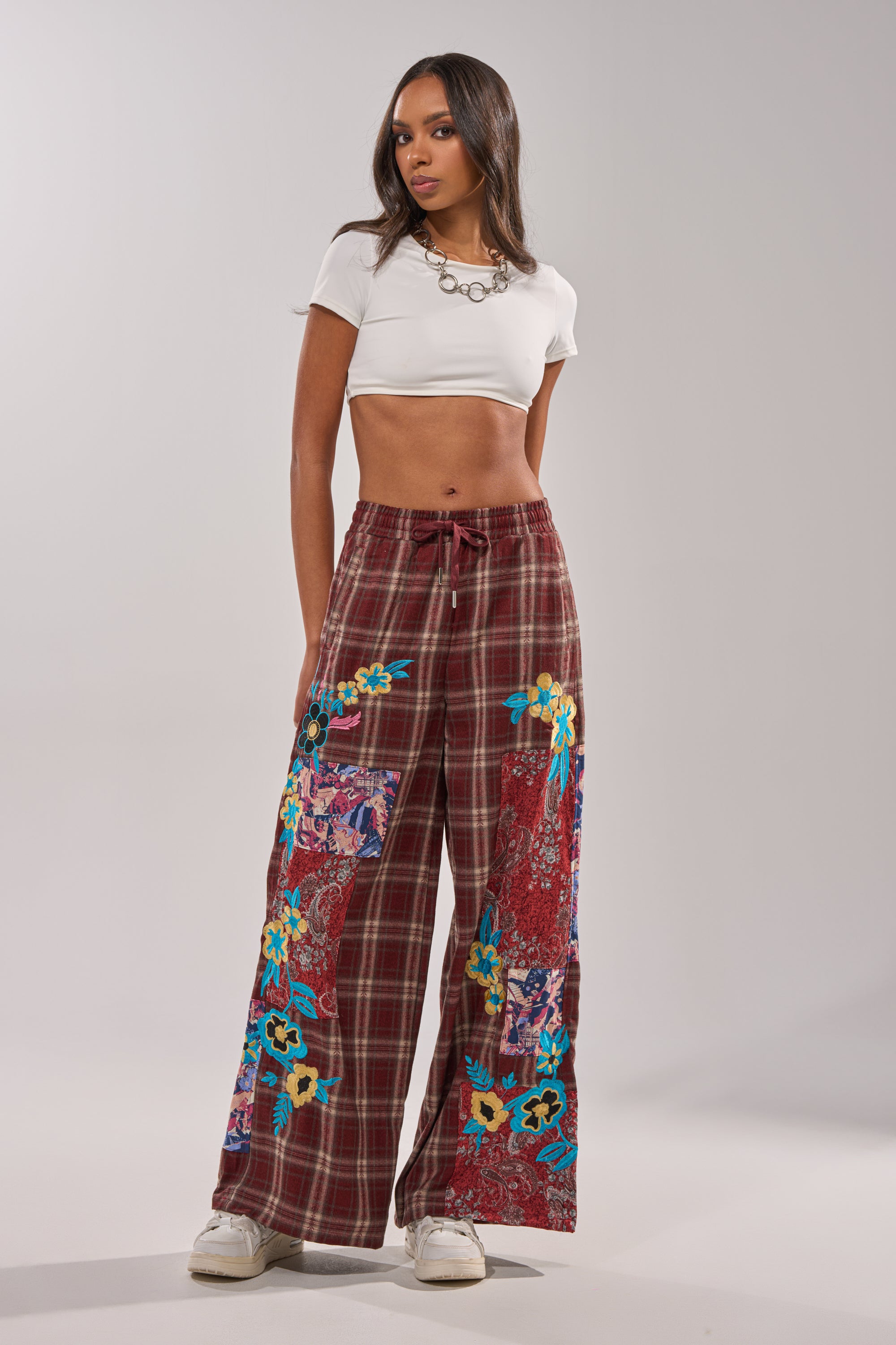 A woman wears the KRIYSTA FLORAL PATCH PLAID PANT with a white crop top and sneakers. She has long hair, a statement necklace, and stands confidently against a plain backdrop. 6